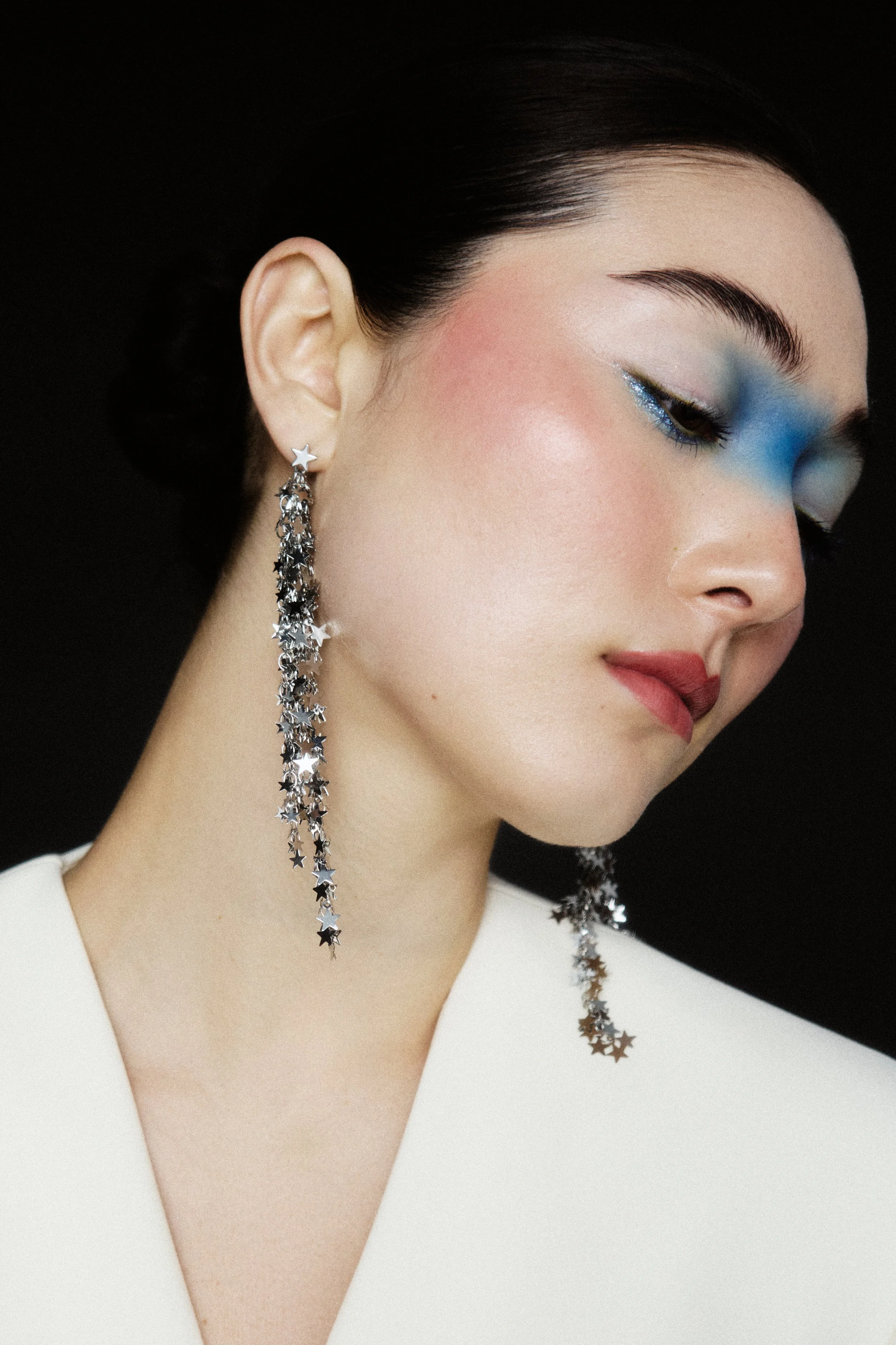 Close-up of a woman's face turned to the side, showing a star-shaped earring with dangling star chains, and makeup featuring blue and silver eyeshadow, blush, and lipstick, against a dark background.