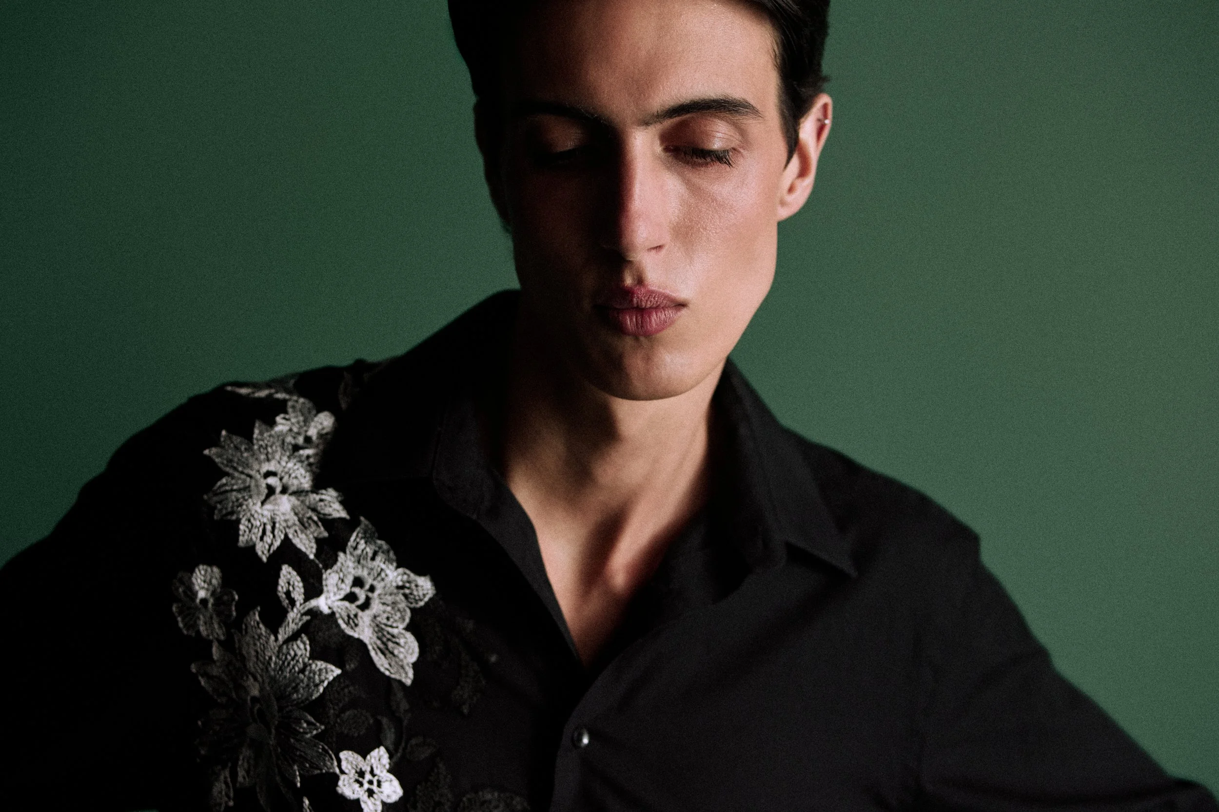 Close-up of a person with short dark hair and closed eyes, wearing a black shirt with embroidered floral patterns on the shoulder, against a dark green background.