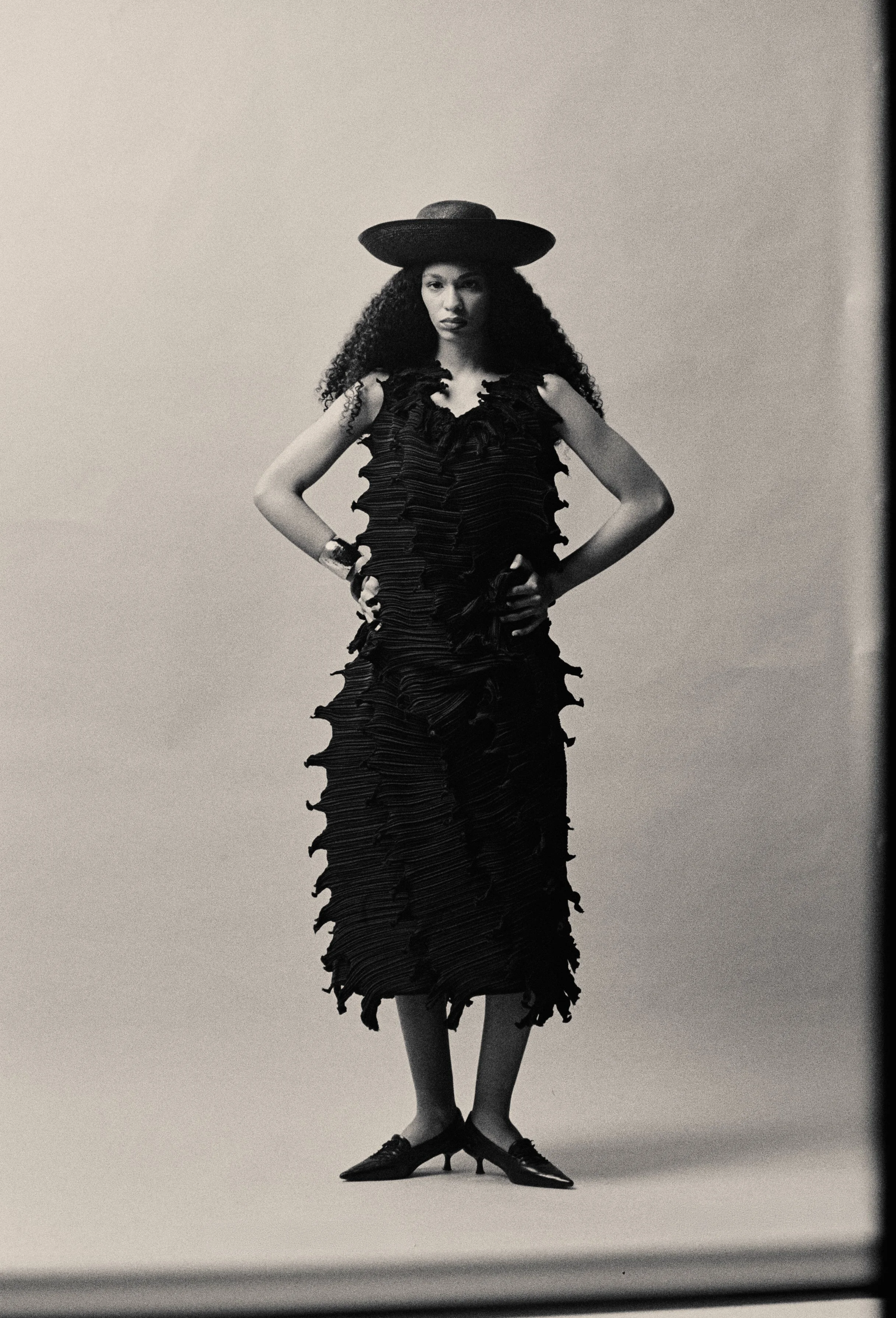 A woman with curly hair wearing a wide-brimmed hat and a ruffled dress stands with her hands on her hips, crossing her legs at the ankles, against a plain background.