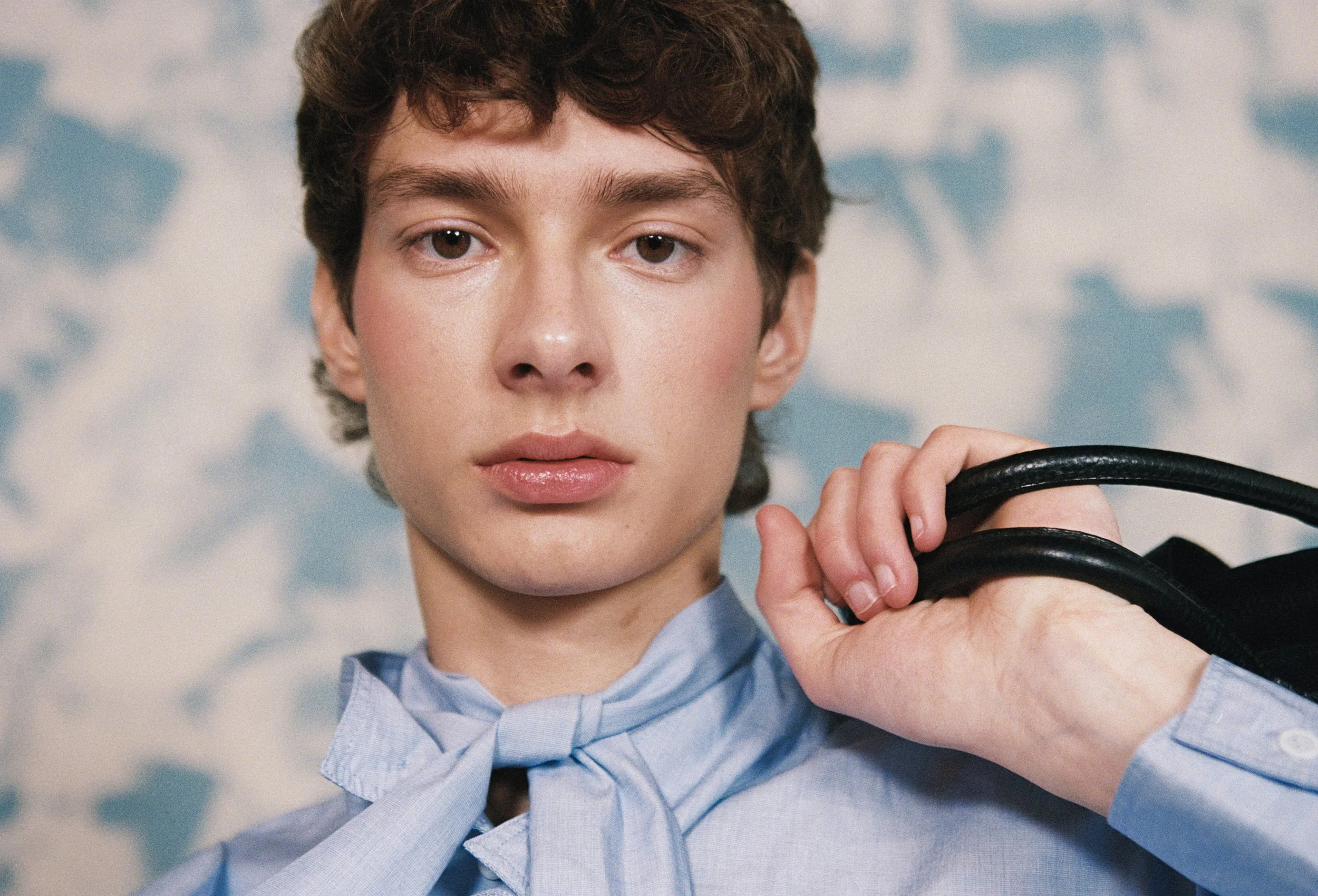 Close-up of a young person with short curly brown hair, wearing a light blue collared shirt with a bow tie, holding a black object with both hands, against a blue and white cloudy sky background.