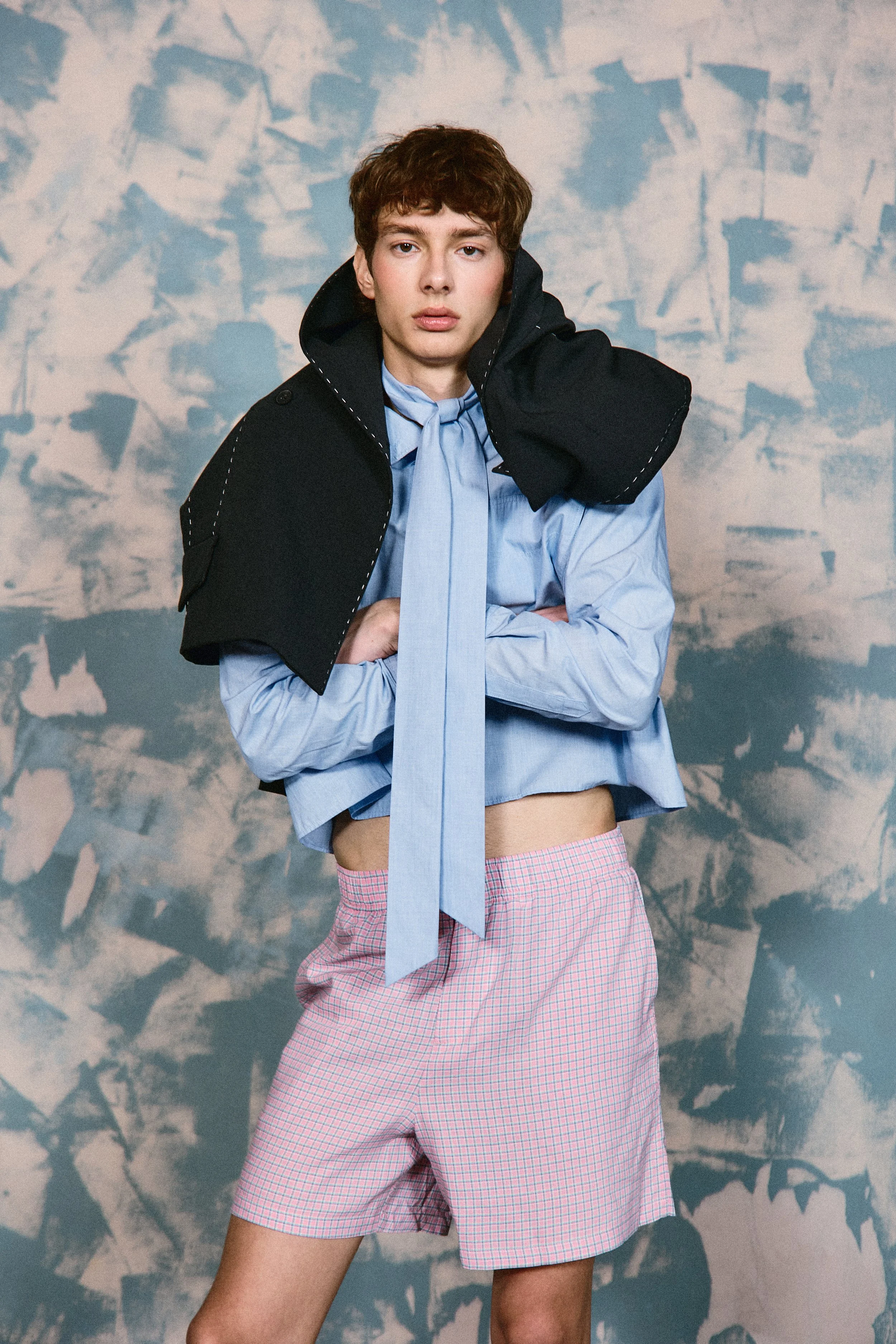 A young person with short brown hair wearing a blue dress shirt, pink checkered shorts, and a black jacket draped over their shoulders, standing against an abstract, blue patterned background.
