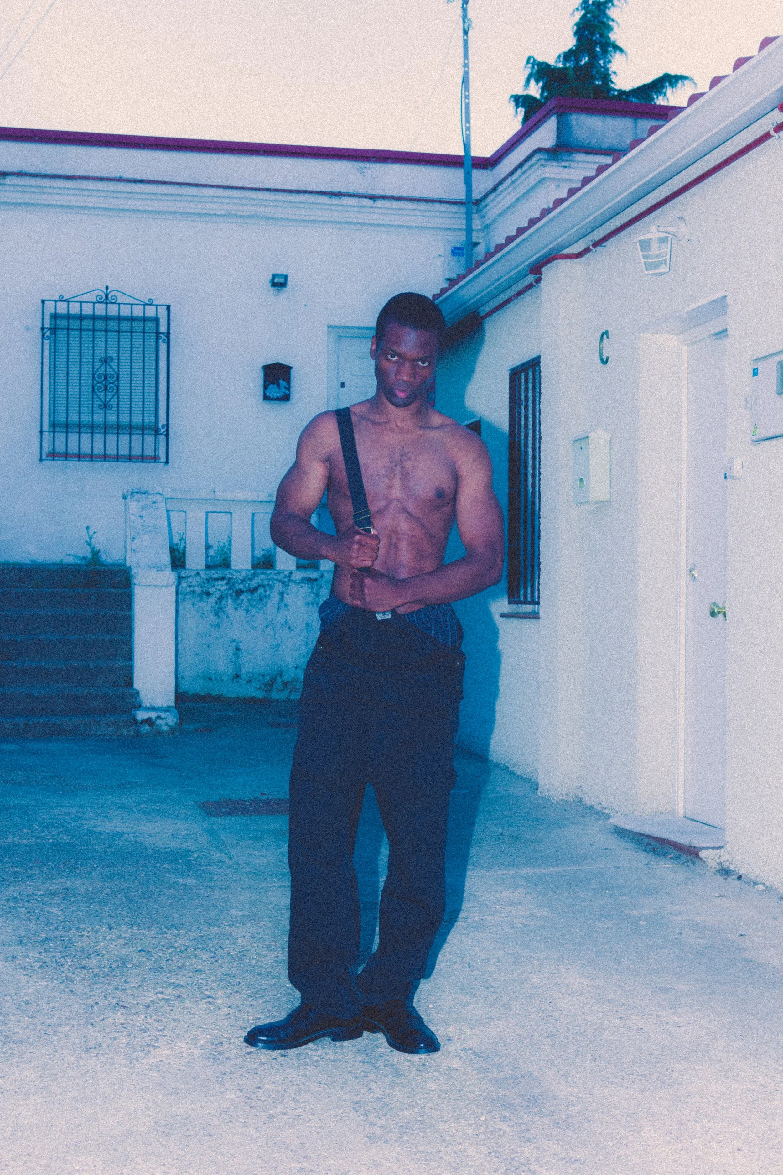 A shirtless man standing outside near a white building with barred windows, wearing dark pants and shoes, holding a strap over his shoulder.