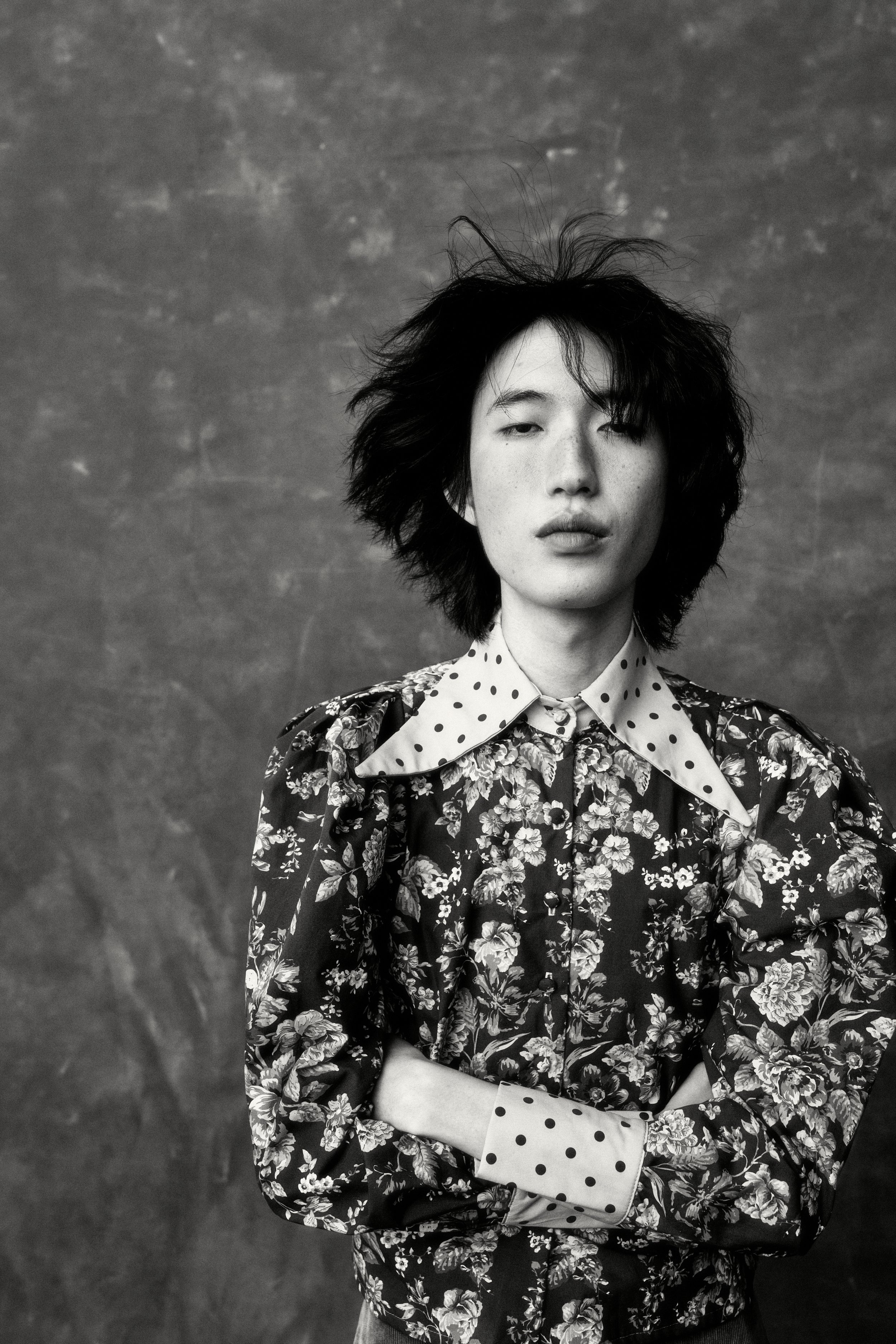 Black and white portrait of a woman with messy hair, wearing a floral patterned shirt with a contrasting polka dot collar and cuffs, standing in front of a textured wall.