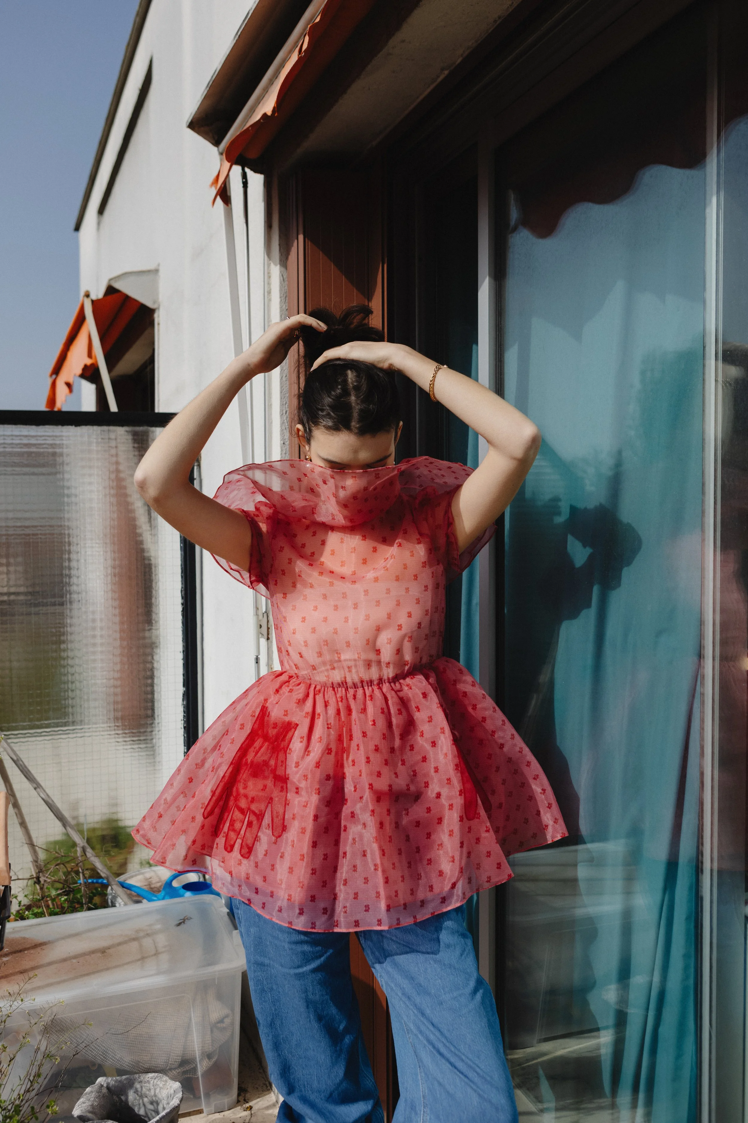 A woman wearing a red, semi-transparent dress with a floral pattern and a tulle skirt is standing on a balcony, tying her hair into a bun. She is wearing blue jeans and is near a sliding glass door and a wooden window.