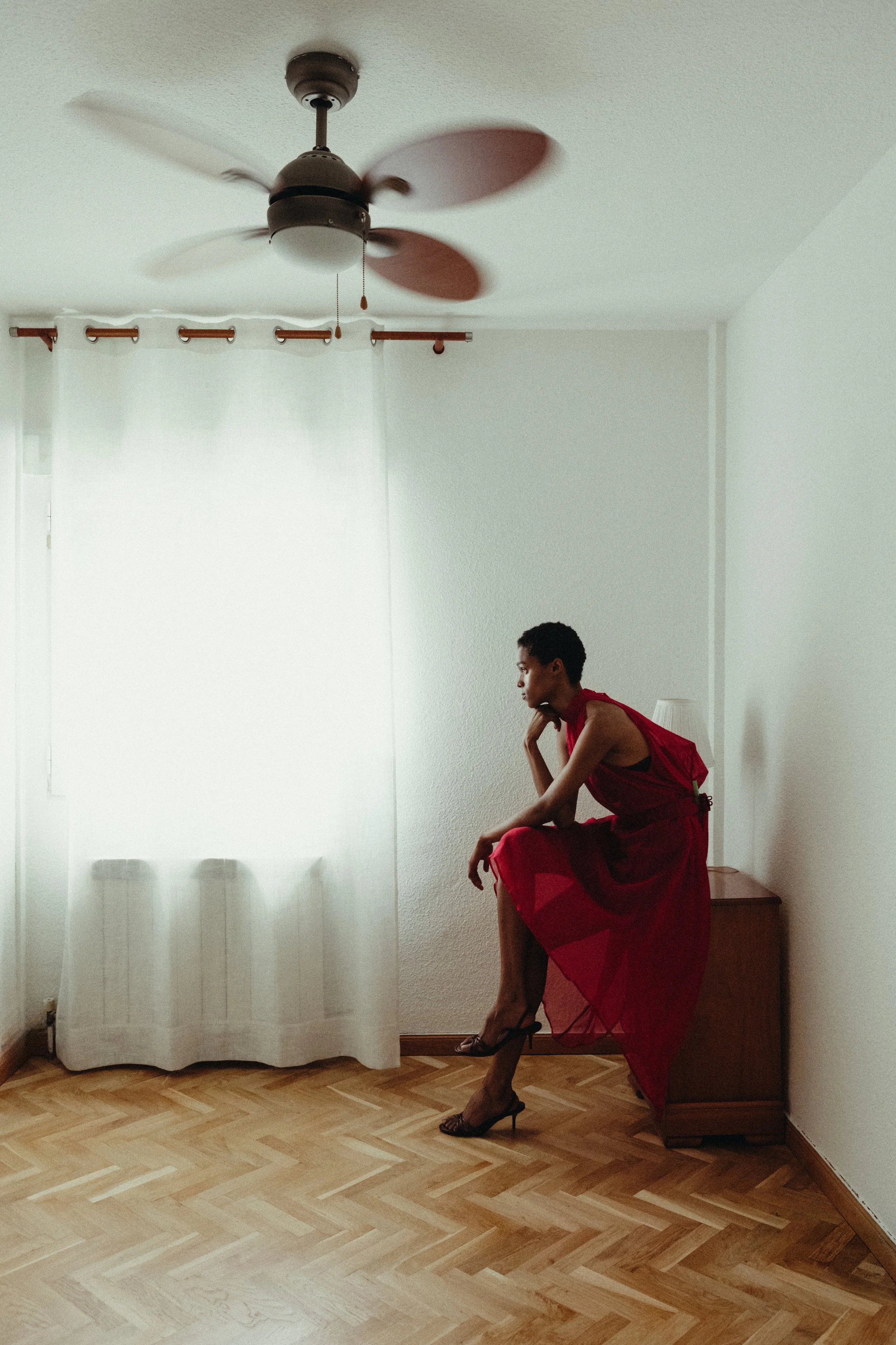 A woman in a red dress and high heels sitting on a wooden dresser in a room with a ceiling fan and white curtains.