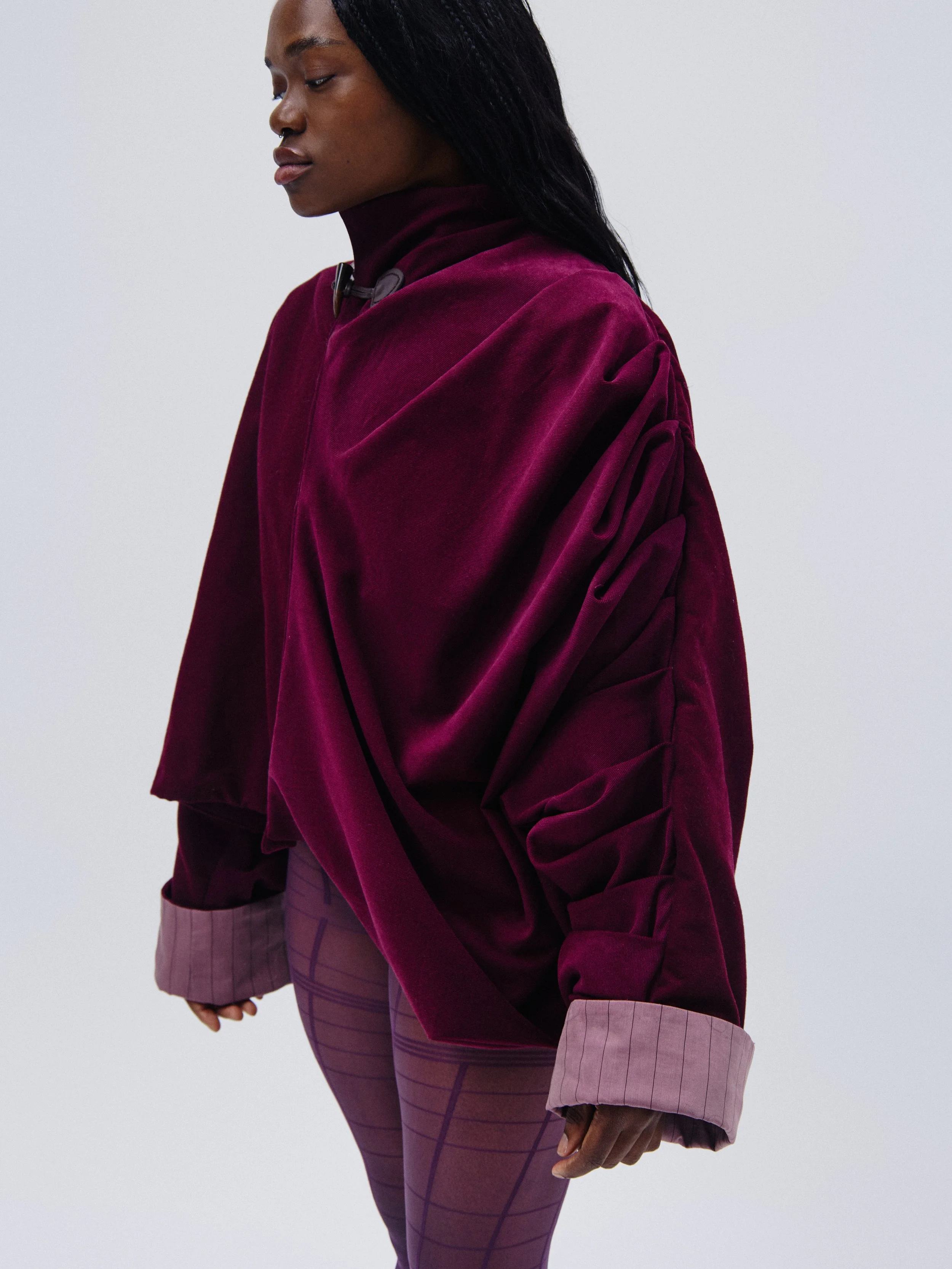 A woman wearing a burgundy velvet jacket with rolled-up sleeves over a matching high-neck top, and purple patterned pants with a grid design, standing against a plain gray background.