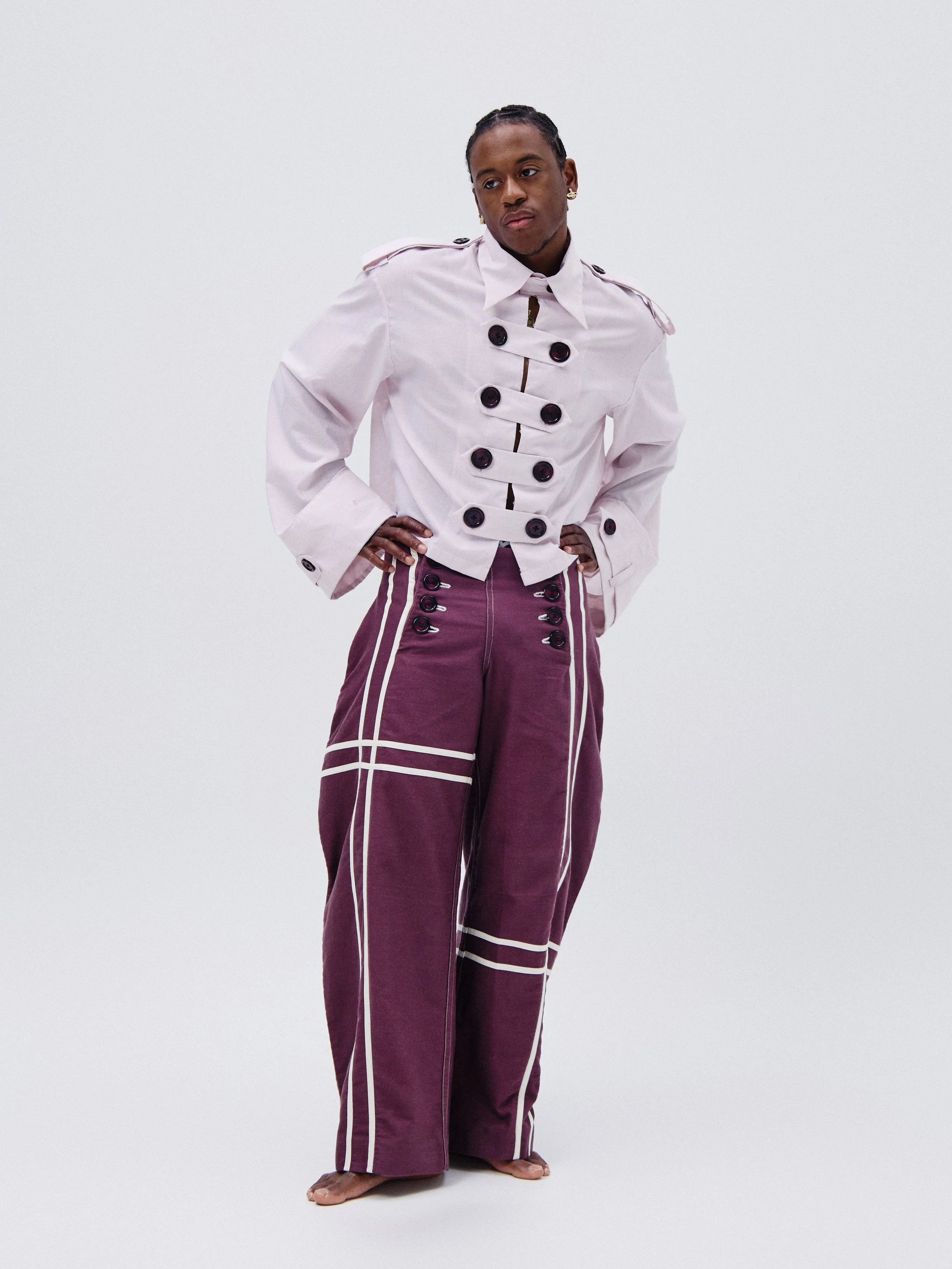 Person standing with hands on hips, wearing a white jacket with black buttons and purple pants with white piping, against a plain white background.
