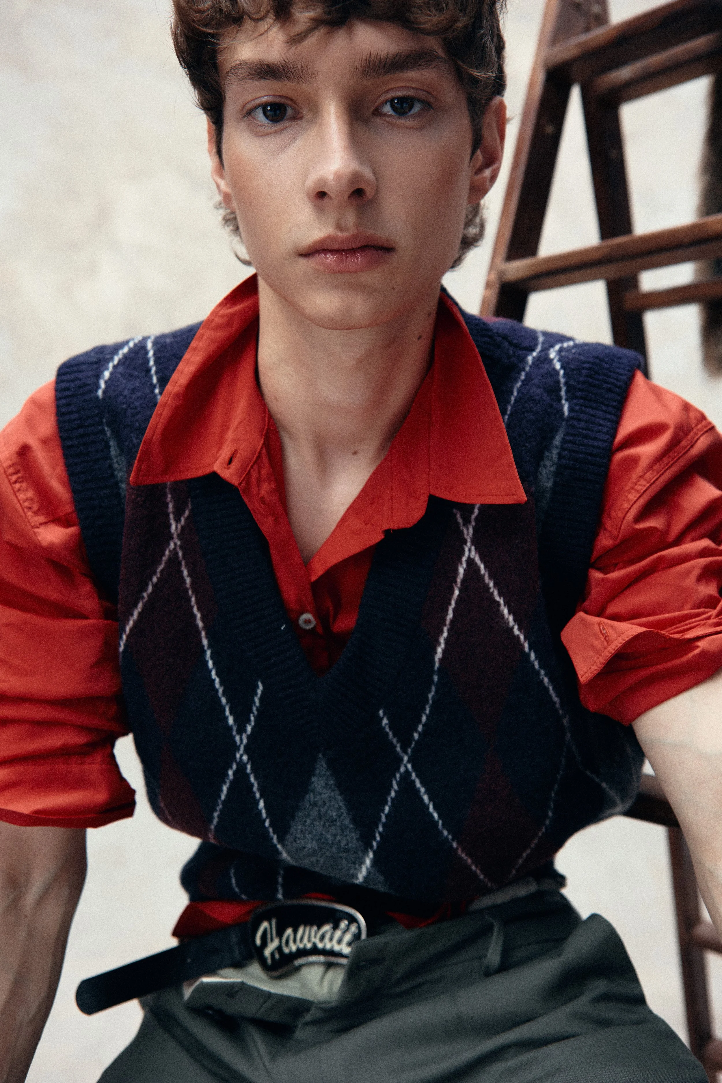 Close-up of a young man with blue eyes and curly brown hair, wearing a red shirt under a dark blue and grey argyle sweater vest, sitting near a wooden ladder.