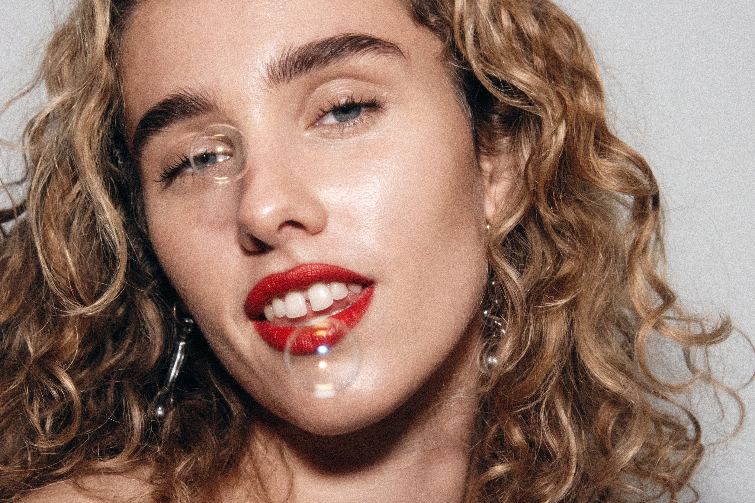 Close-up of a woman's face with curly hair, red lipstick, and pearl earrings, smiling and blowing bubbles