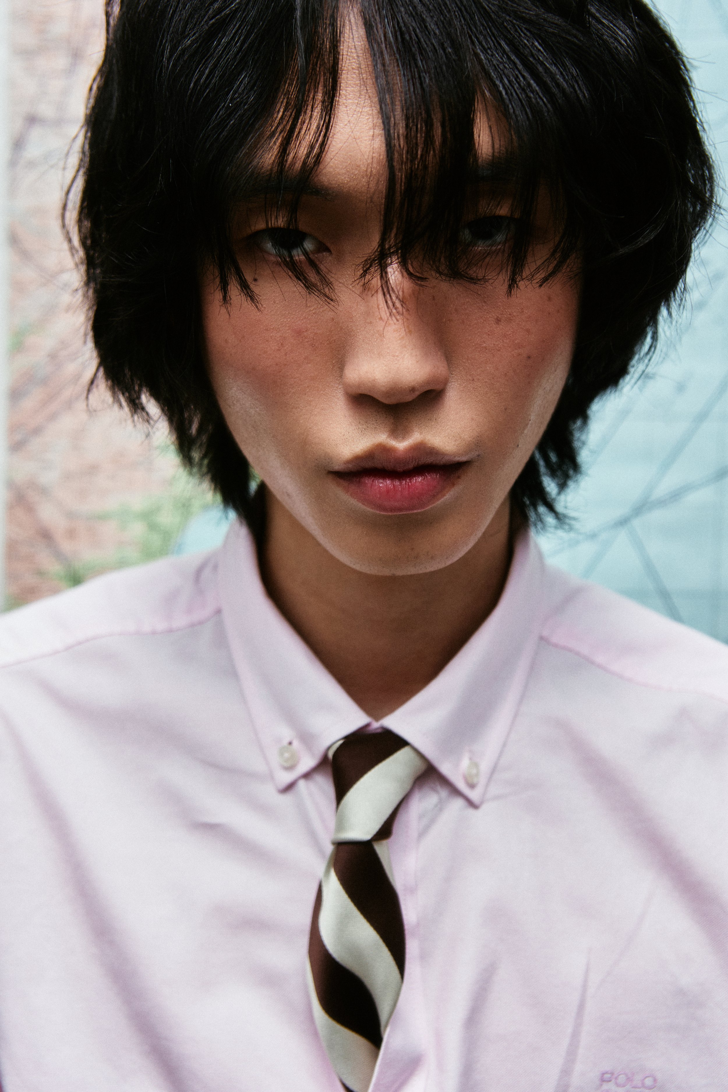 Close-up of a young person with short black hair, wearing a light pink collared shirt and a striped tie, against a blurred outdoor background.