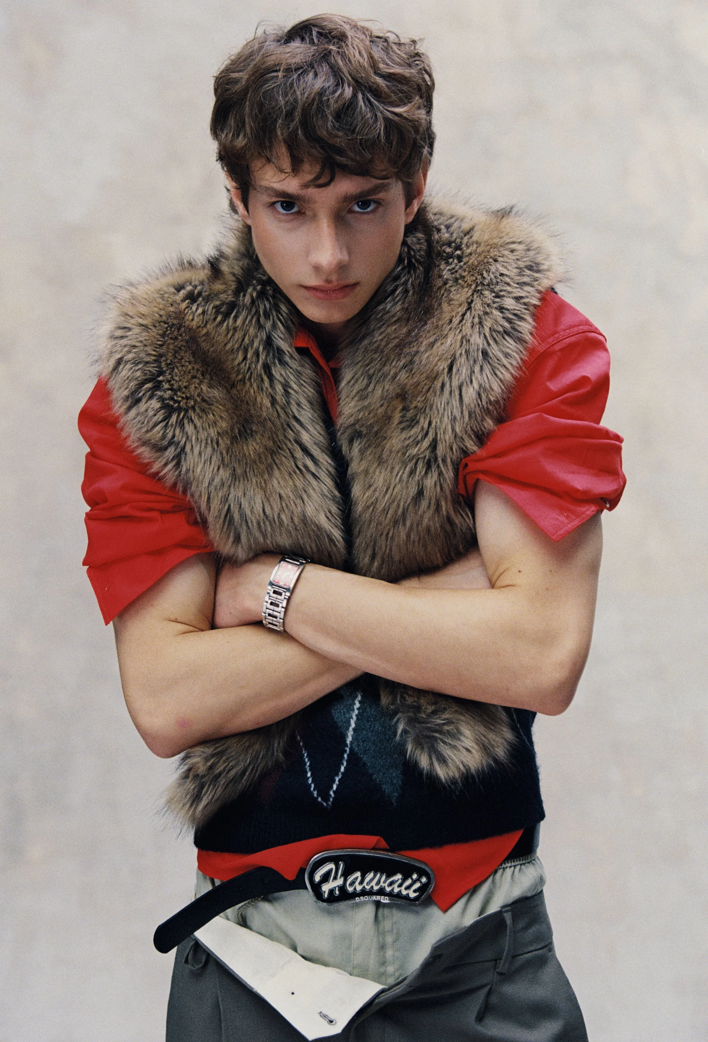 A young man with brown, curly hair and blue eyes looking directly at the camera. He is wearing a red shirt with rolled sleeves, a fur vest, and has a serious expression. His arms are crossed, and he is standing with a neutral background. He also has 