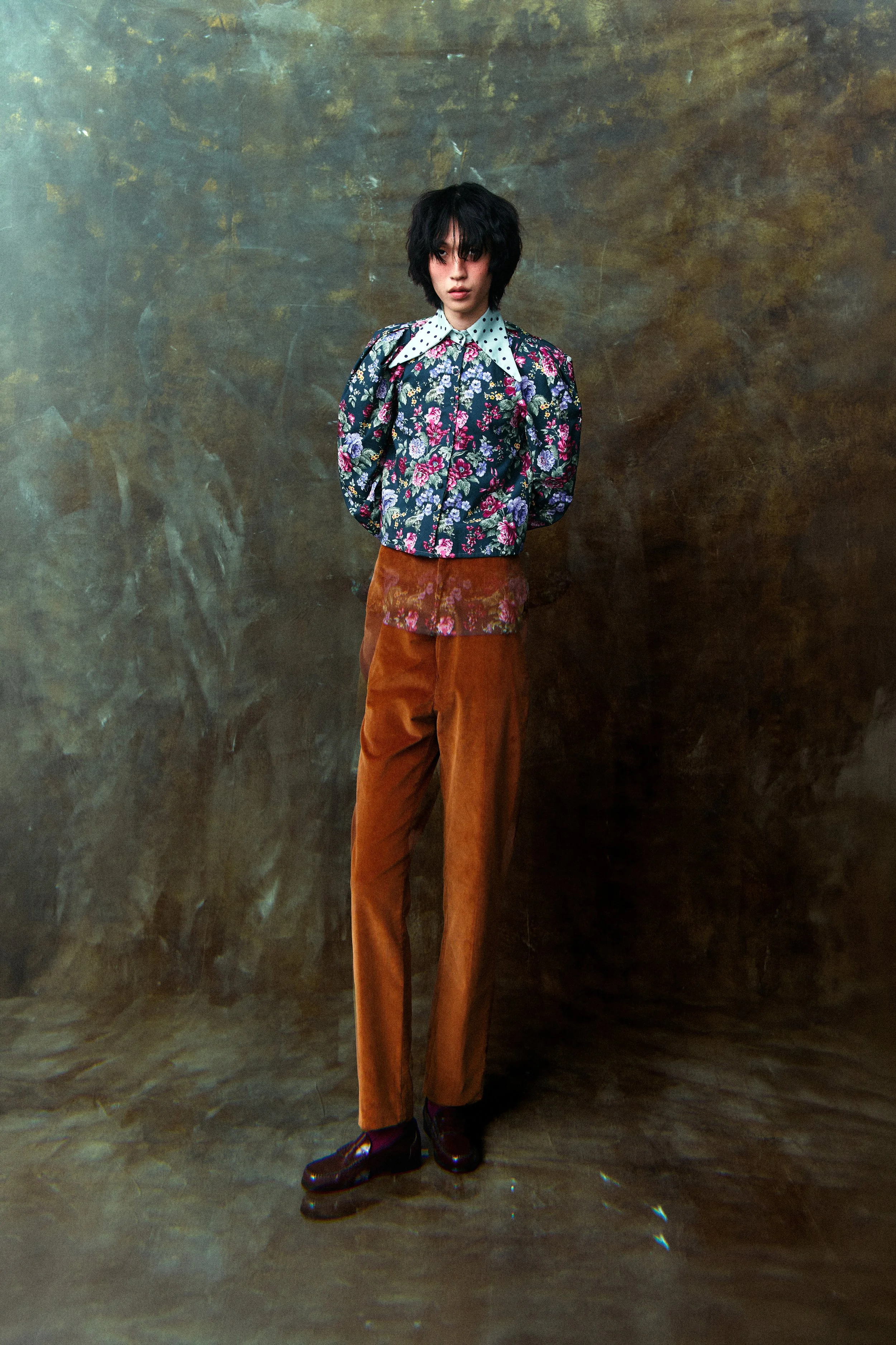 A young person with dark, messy hair standing against a textured, brownish background. They are wearing a floral patterned jacket over a collared shirt with polka dots, loose brown trousers, and dark shoes. Their expression is neutral, and they are l