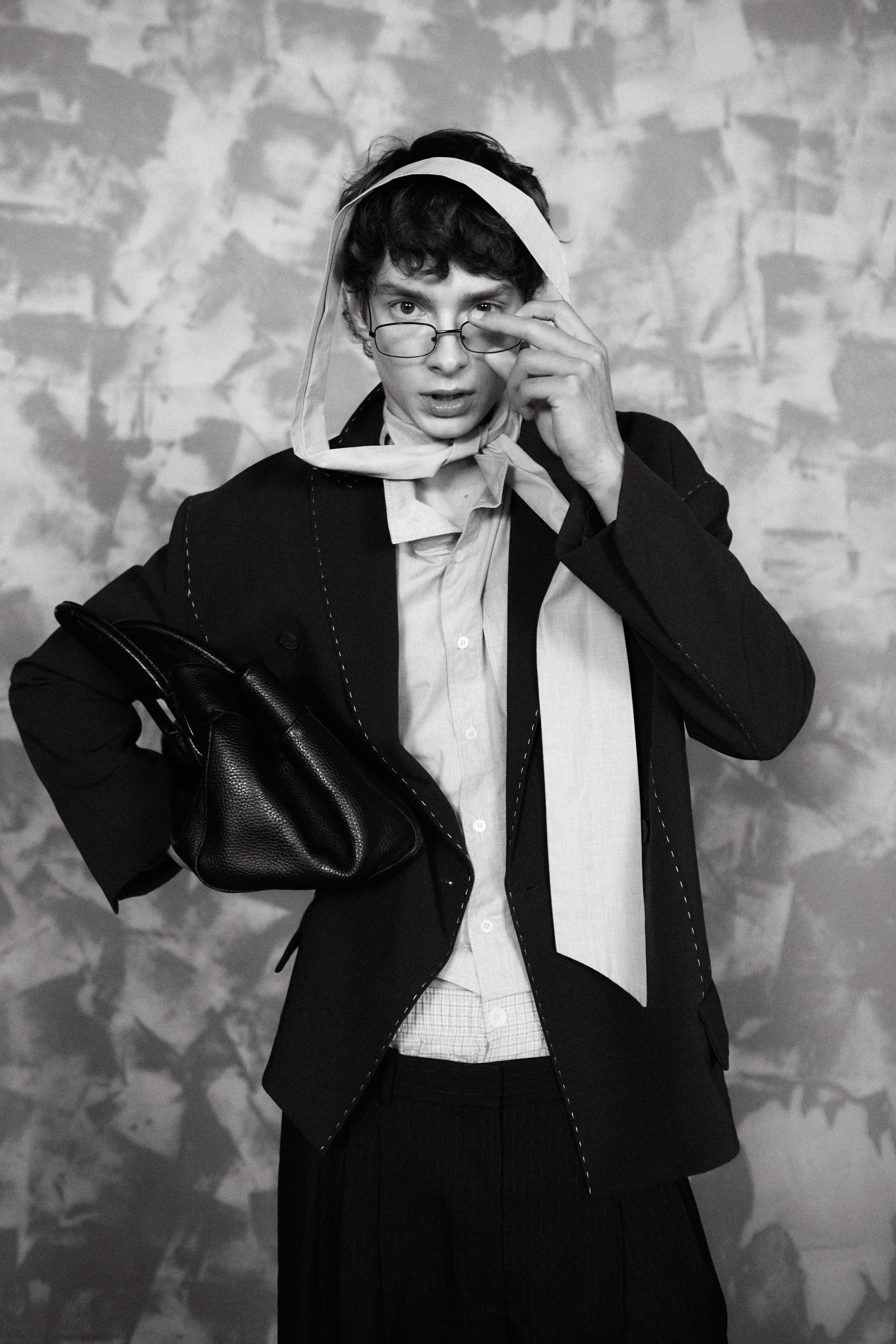 A young person with curly hair wearing glasses, a coat, and a scarf, holding a bag, standing against a textured background.