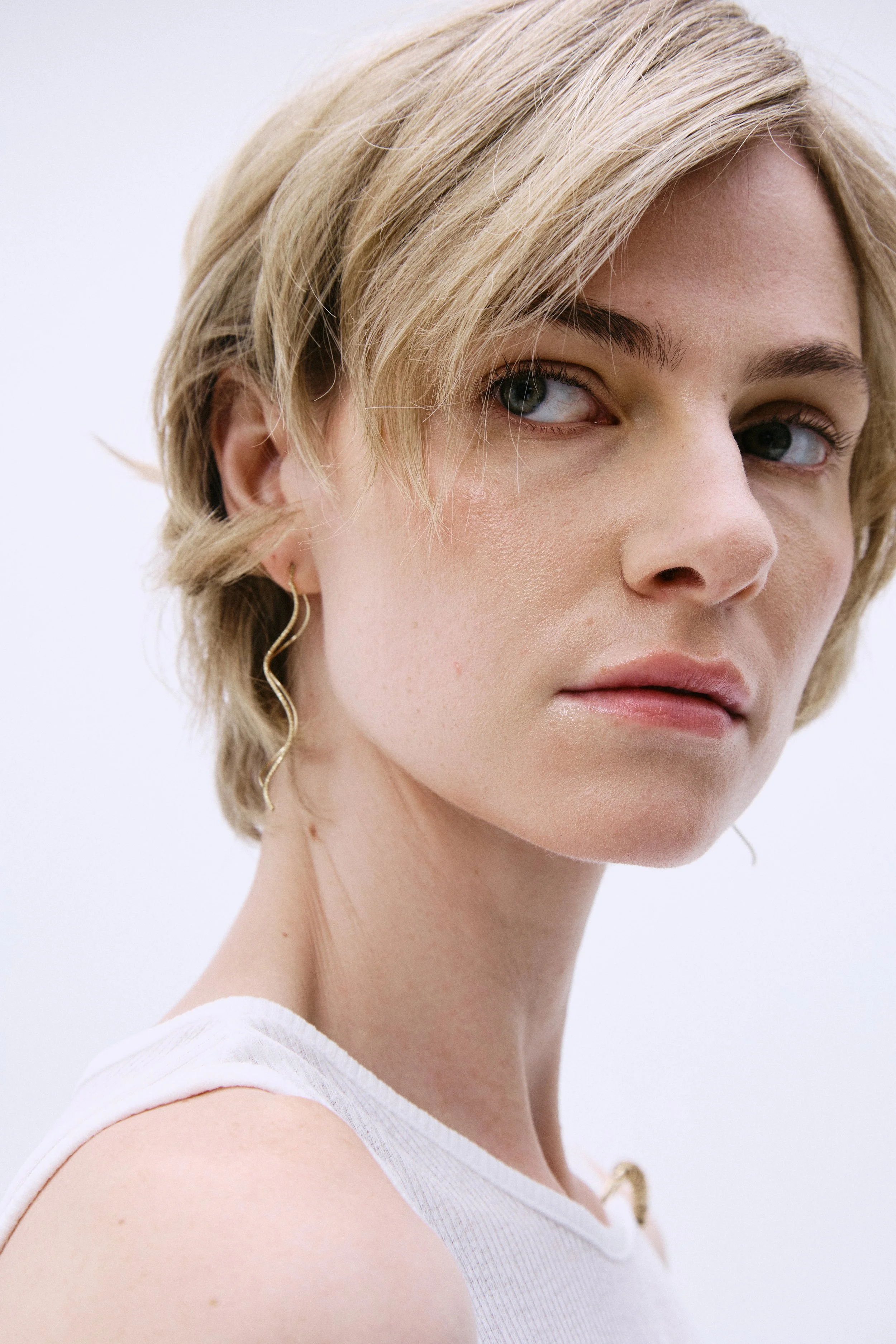 Close-up of a blonde woman with short hair, wearing a white top and gold earrings, looking to the side against a plain light background.