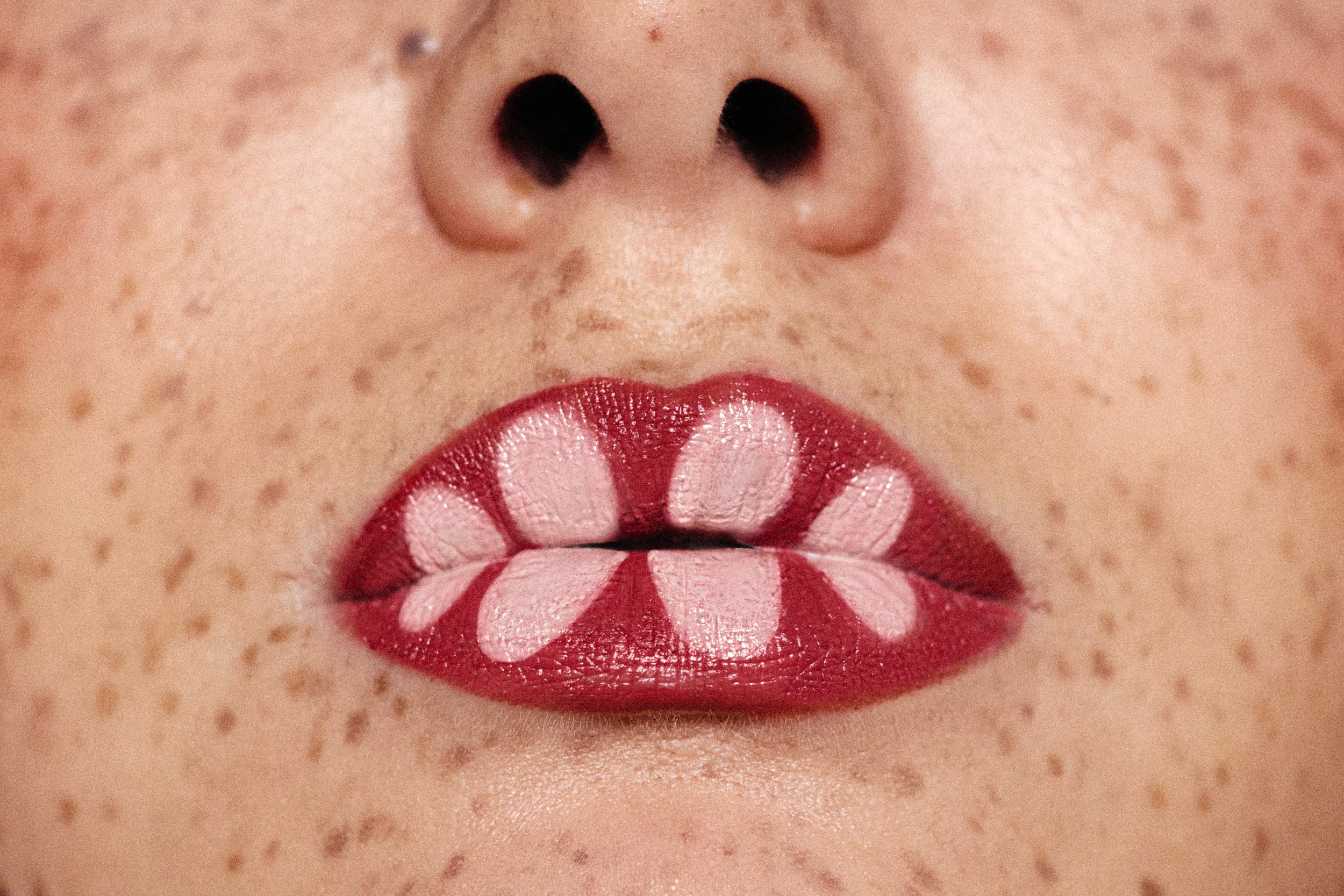 Close-up of a person's lips decorated with red, white, and black lipstick forming a butterfly pattern, with freckles and skin texture visible.
