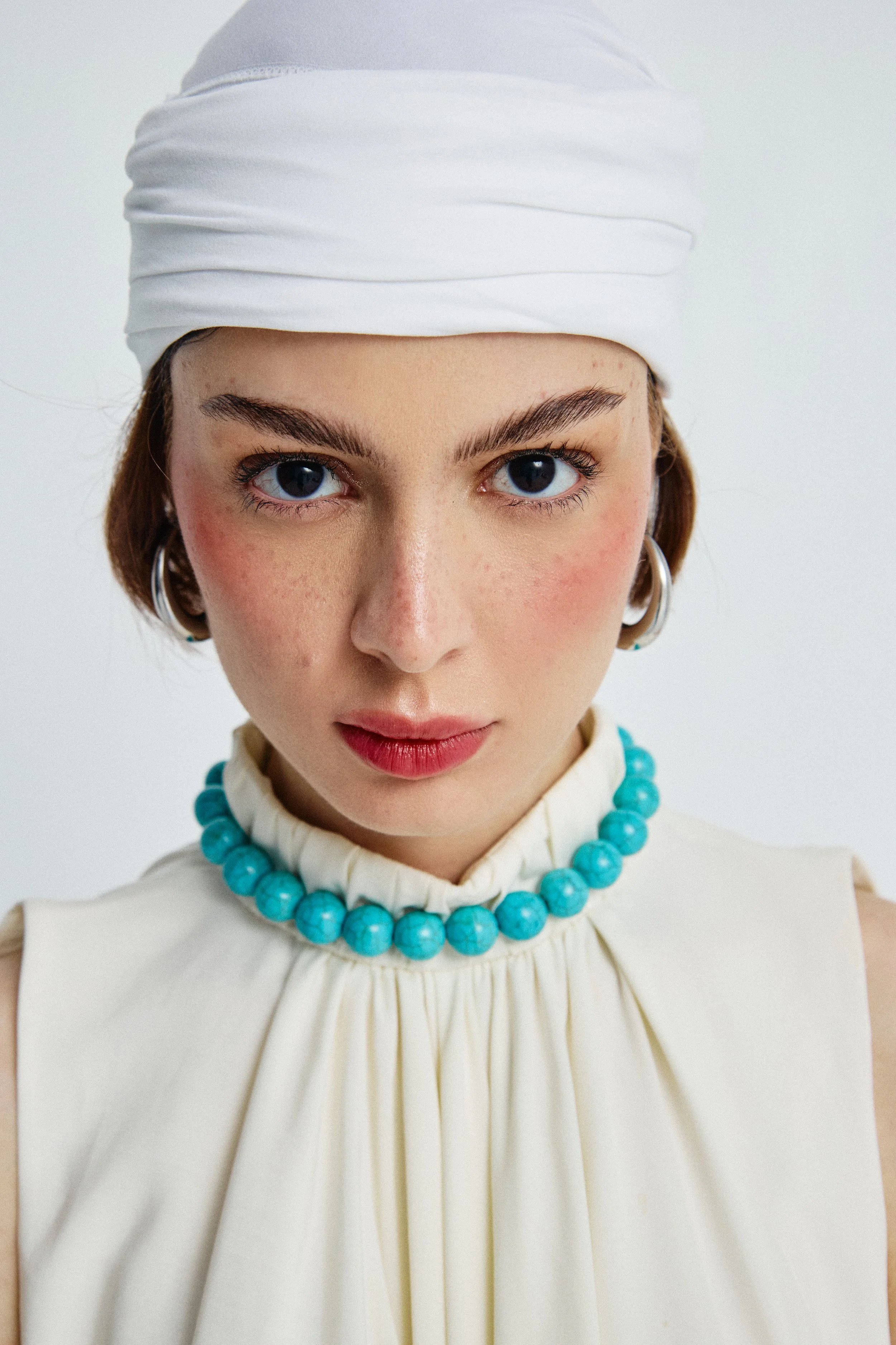 Close-up of a woman wearing a white head wrap, large turquoise beaded necklace, earrings, and a high-neck white dress with pleats.