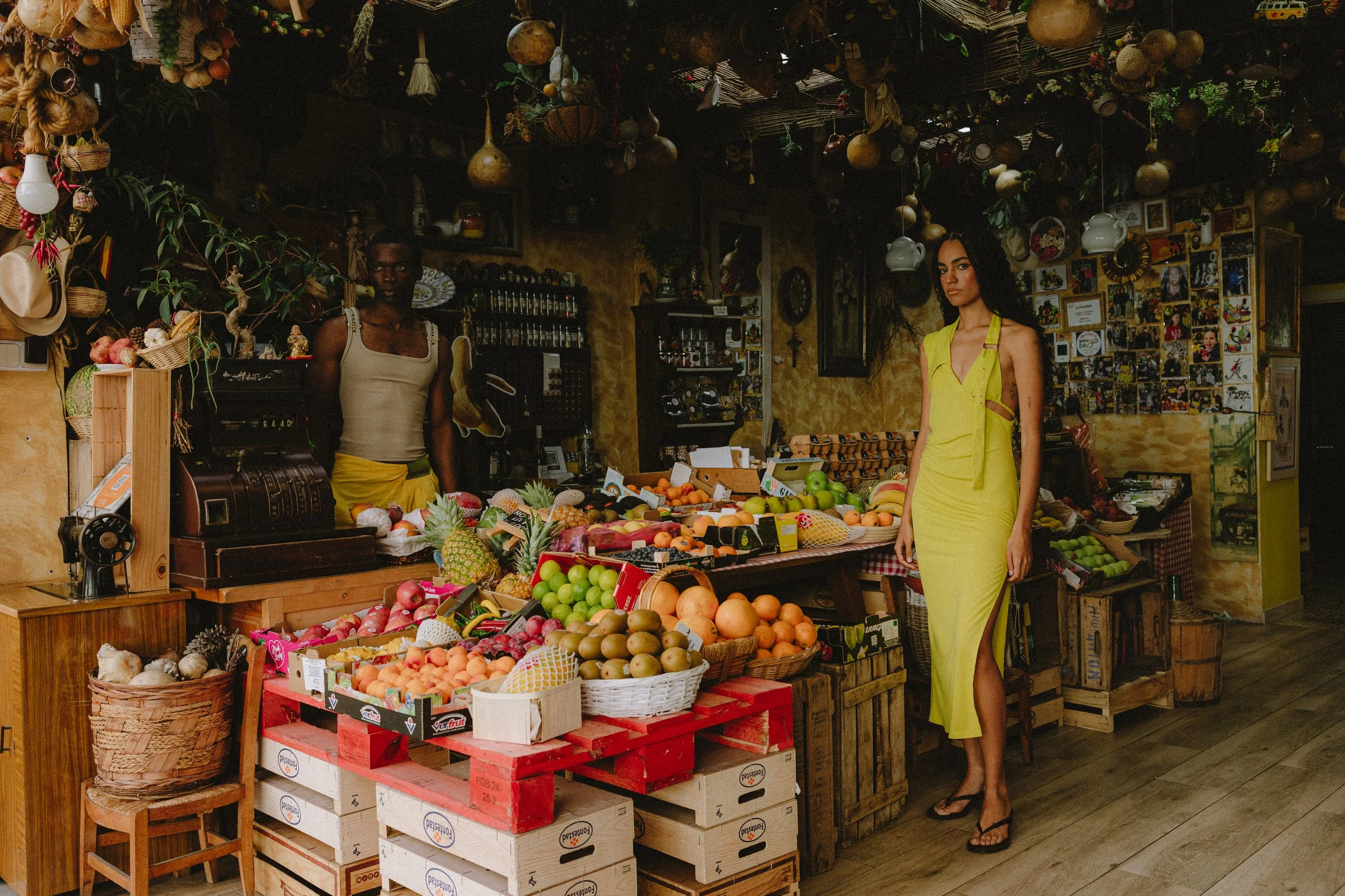 Two women shopping at a produce stand filled with fresh fruits and vegetables. One woman is wearing a yellow dress and the other is in a beige top and yellow skirt. The stand has hanging plants and various framed pictures on the wall behind.