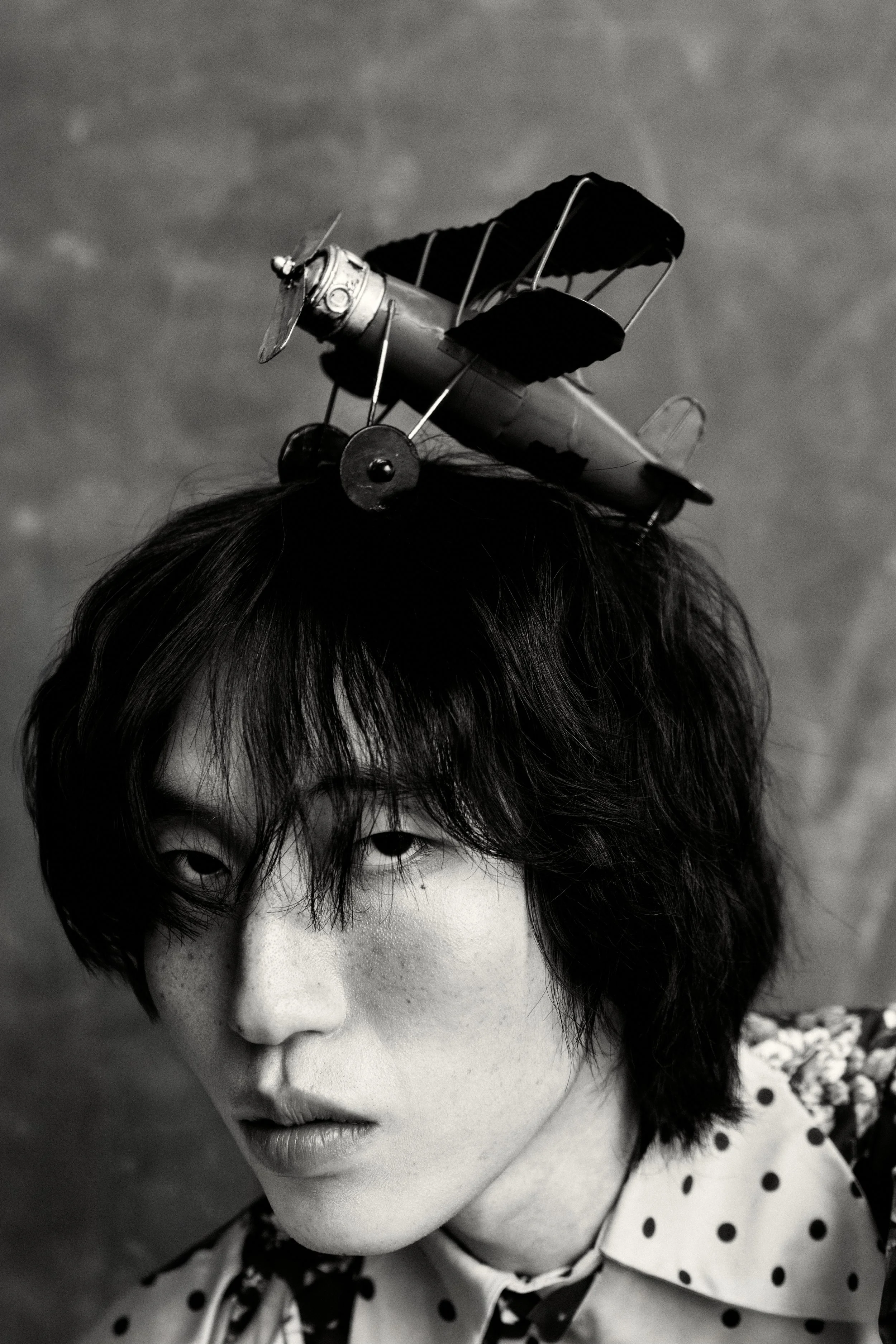 A black and white photo of a young person with dark hair and freckles, wearing a polka-dotted collared shirt. An toy airplane with dark fabric wings is balanced on their head.