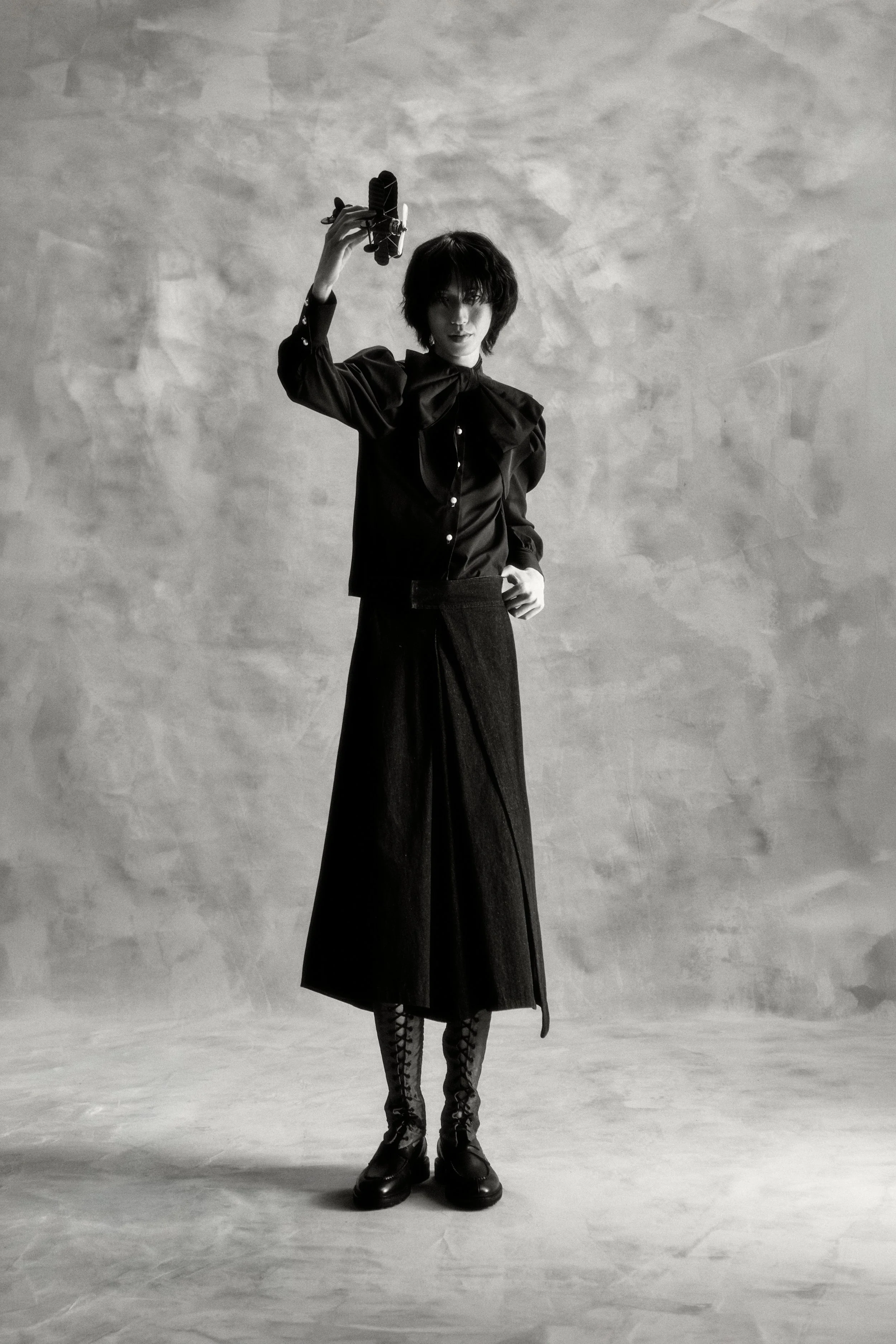 A woman with short, dark hair wearing a long, dark skirt, lace-up boots, and a dark blouse with puffed sleeves, standing against a textured gray wall. She is holding a small vintage-style airplane模型 in the air with her right hand.