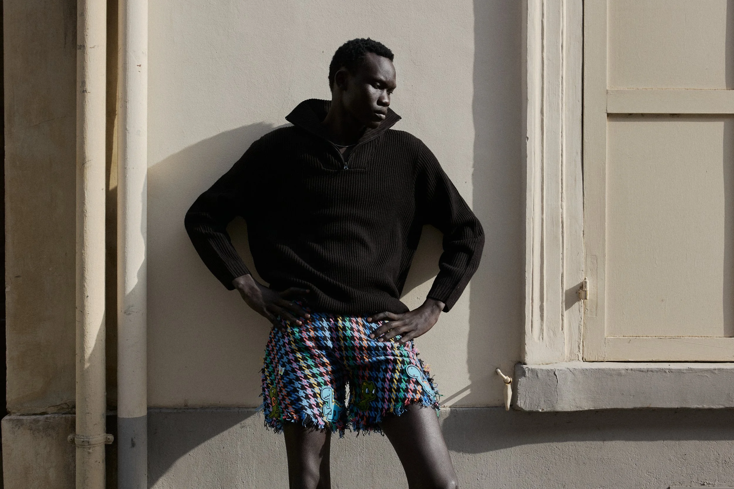 A young Black person standing against a beige wall with a window, wearing a black sweater and colorful, patterned shorts, with hands on hips, looking down and away.