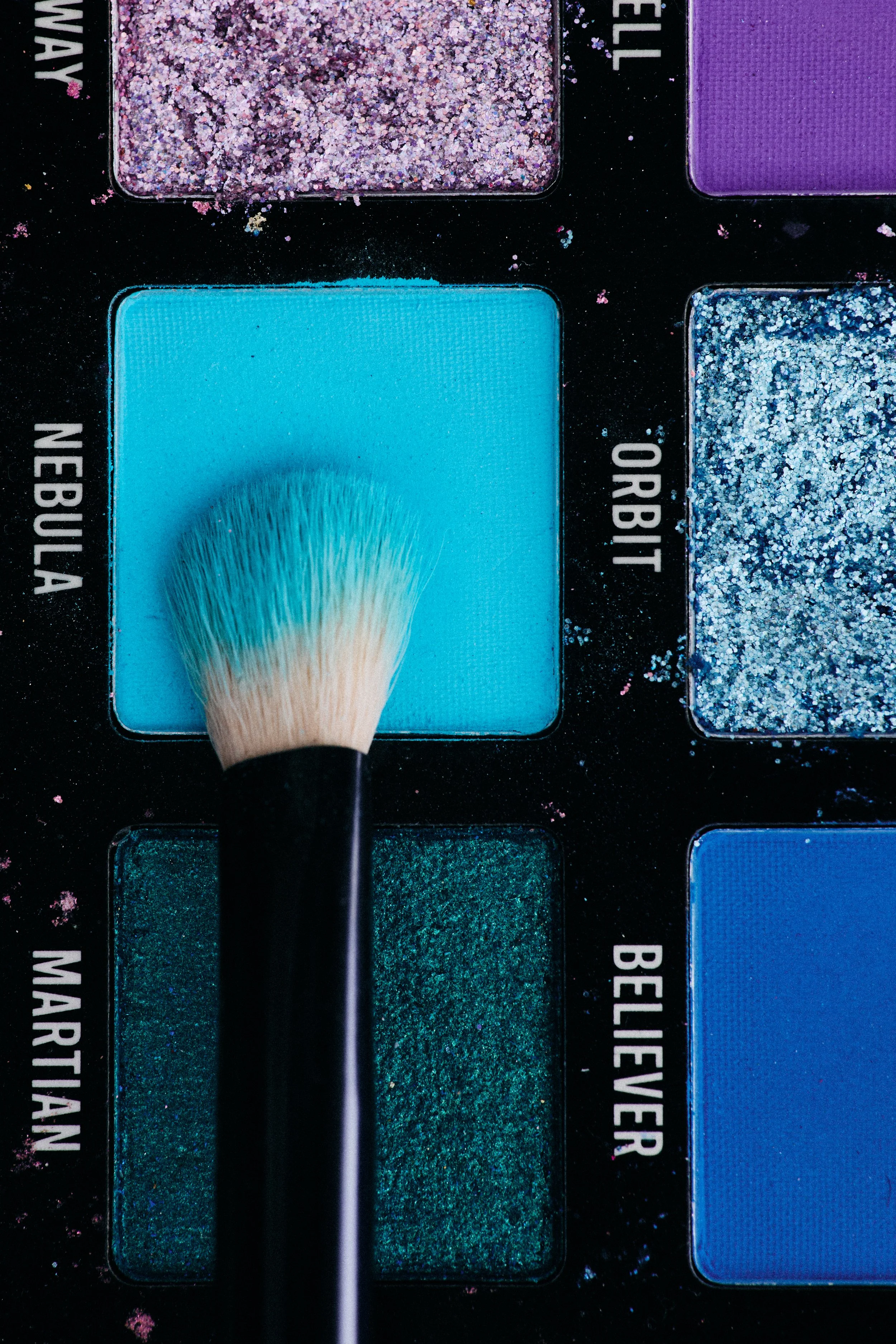 A makeup brush is pressing onto blue eyeshadow in a makeup palette with various shades of blue, green, purple, and glitter colors surrounding it.