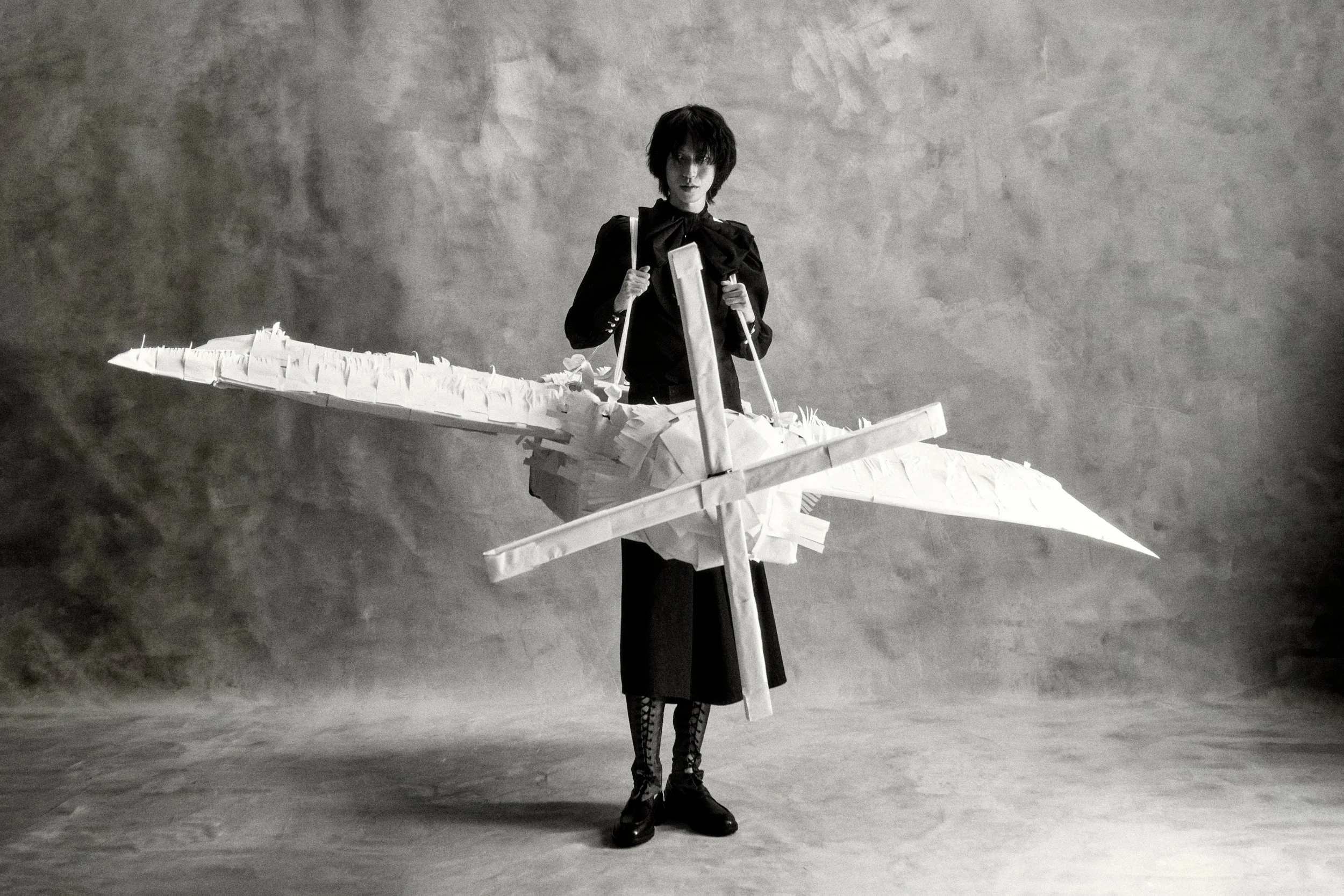 A person with short dark hair stands on a plain floor and holds a large paper or cardboard model of a flying aircraft or spacecraft, with large wings and a tail. The person is dressed in black with patterned socks and boots, and looks directly at the