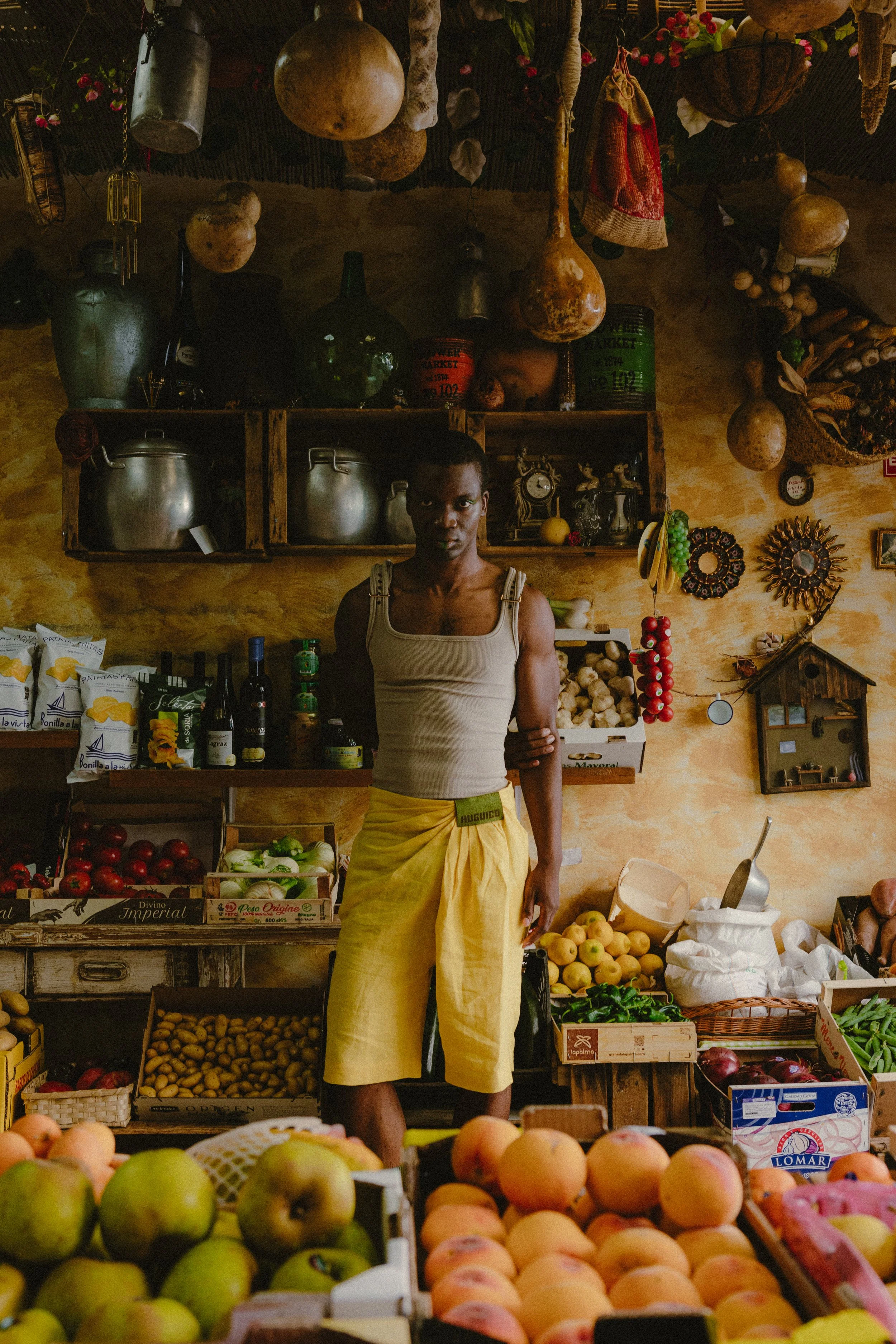 A person standing behind a produce stand filled with apples, potatoes, and other fruits and vegetables at a market, with hanging gourds and decorative items on the wall behind.