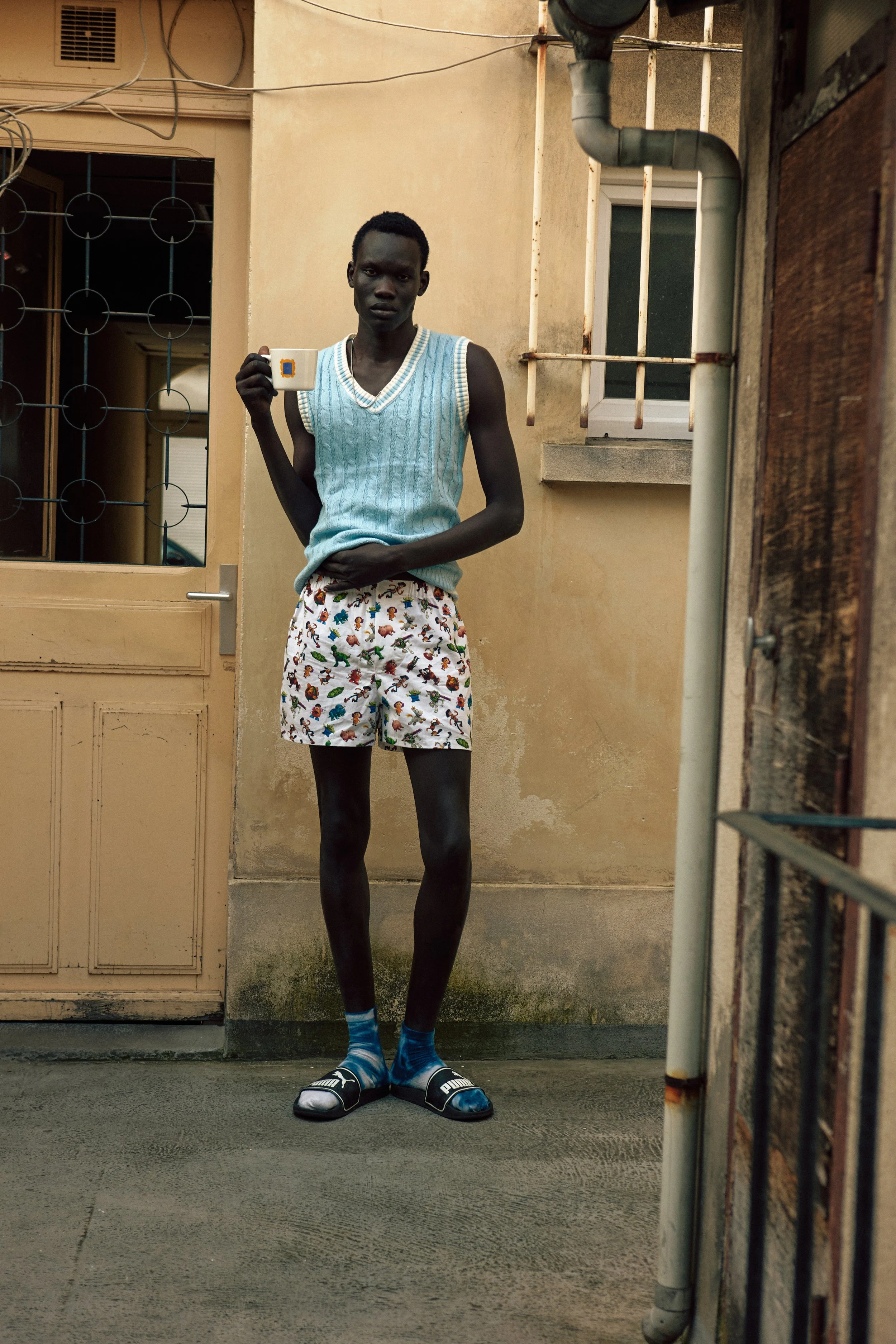 Young man standing outdoors in front of a beige building, wearing a blue sleeveless sweater, animal print shorts, and socks with sandals, holding a white mug.