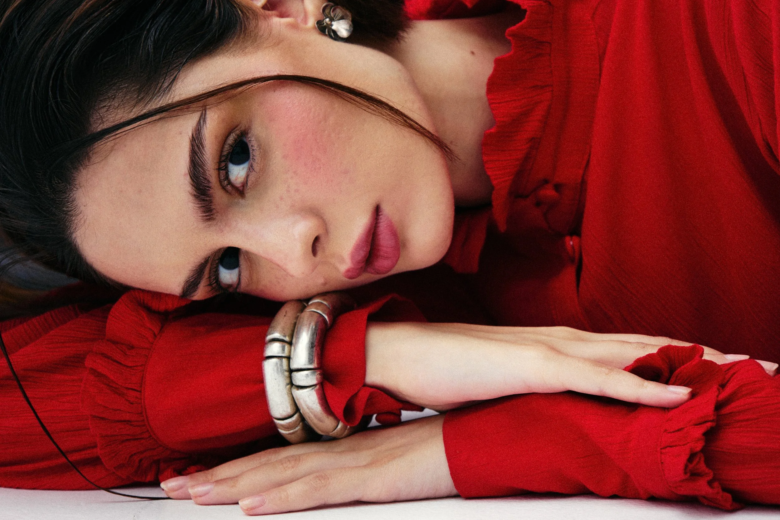 Close-up of a woman with dark hair lying on a white surface, wearing a red ruffled top and silver jewelry, with her head resting on folded arms.