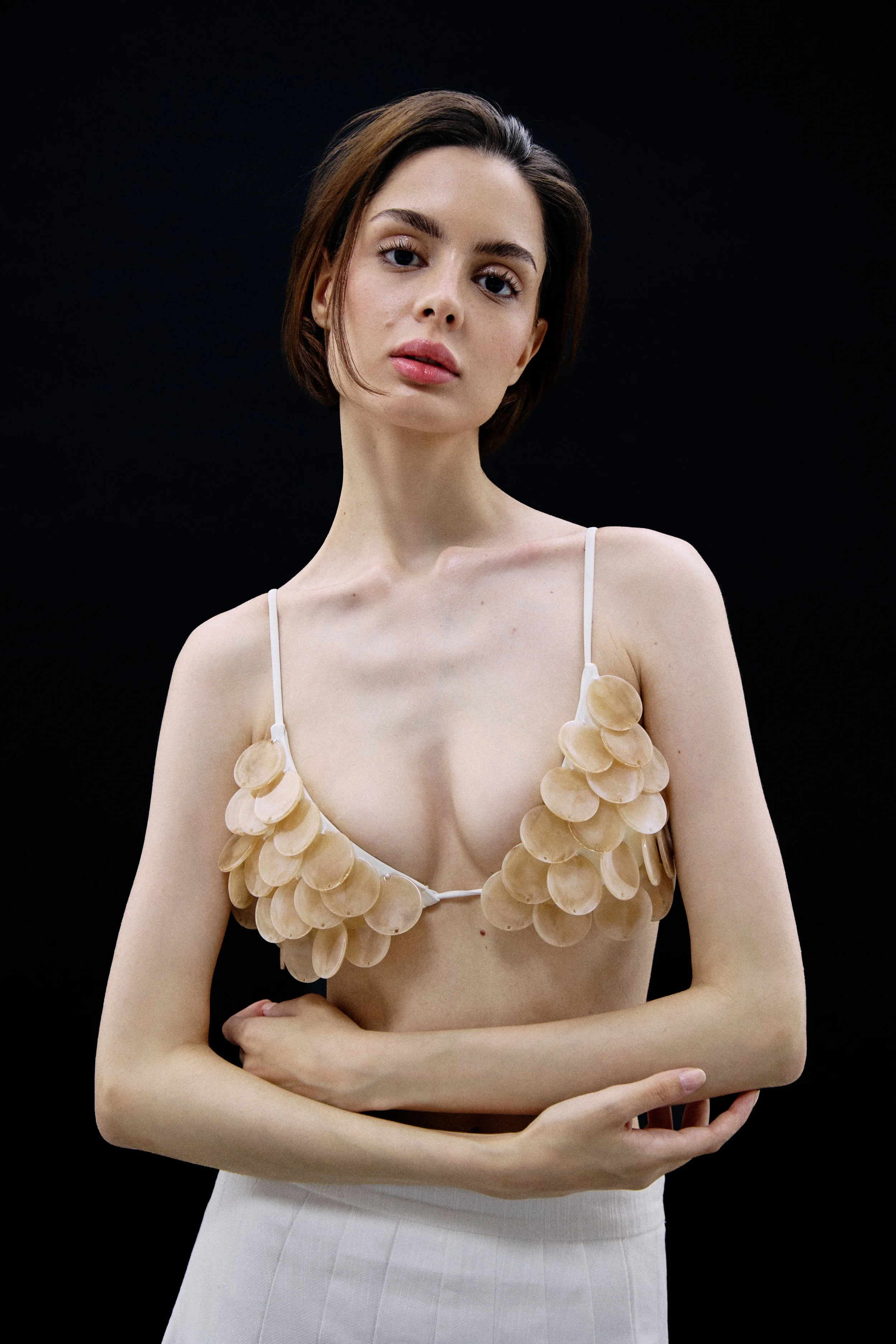 A woman with short brown hair and fair skin wearing a beige top with large oval embellishments and a white skirt against a black background.