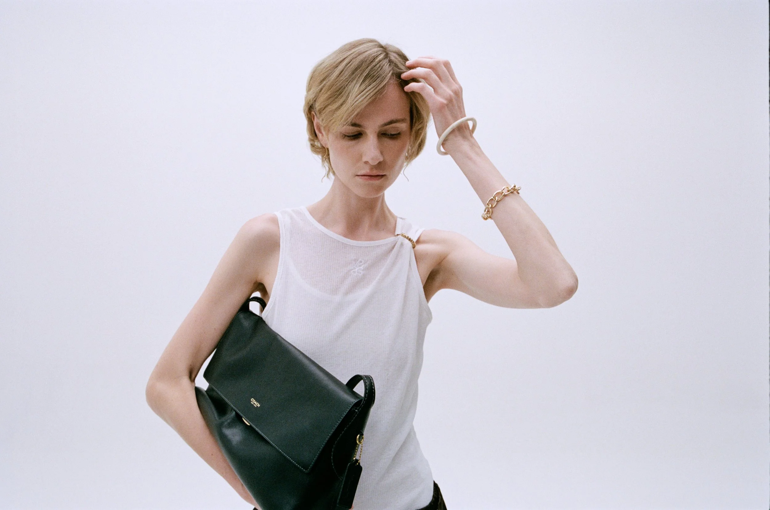 A woman with short blonde hair wearing a sleeveless white top, accessorized with a gold bracelet and a white bracelet, carrying a black handbag with a gold zipper, standing against a plain white background.