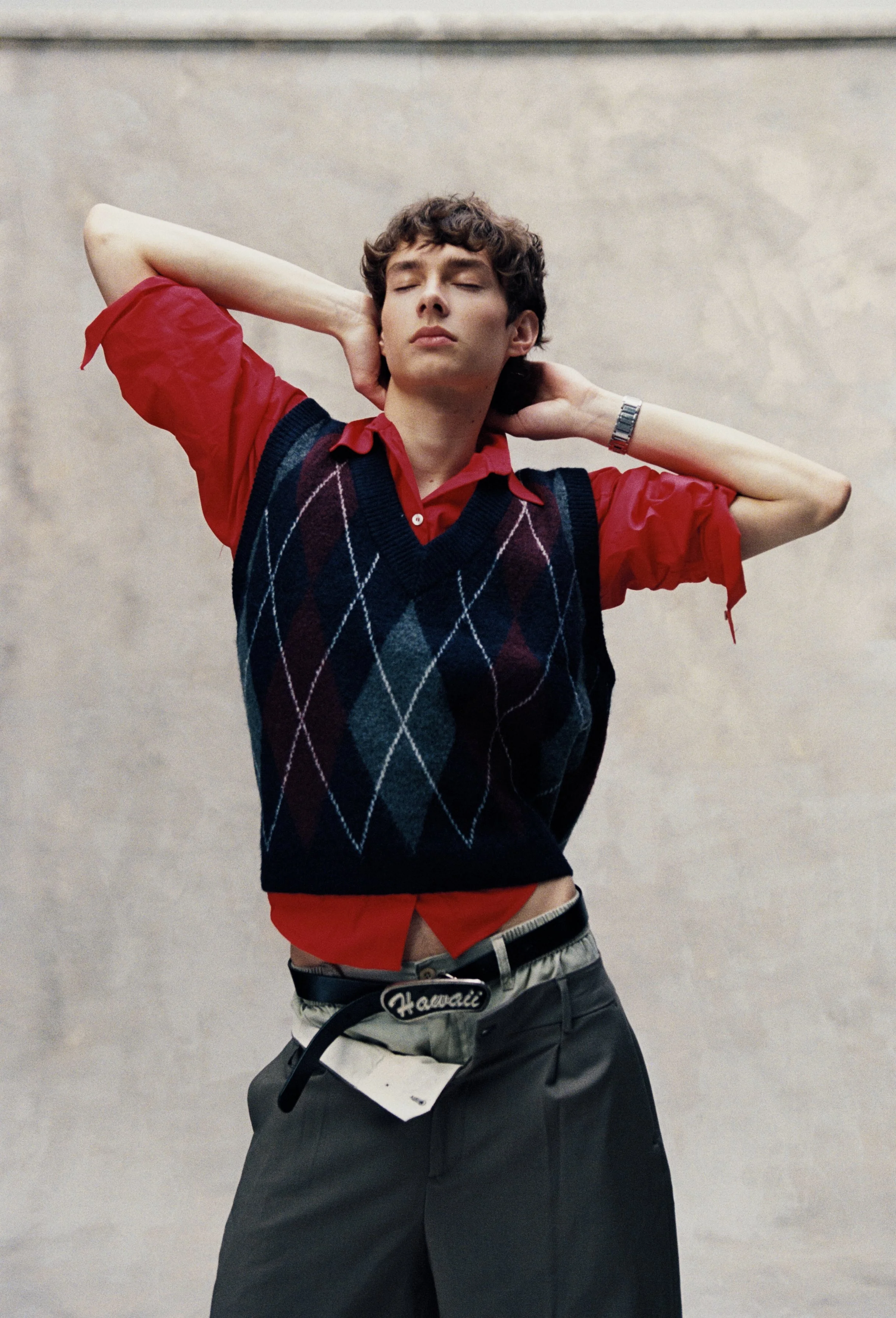 A young man with curly brown hair poses with eyes closed, wearing a red shirt with a navy and maroon argyle sweater vest, gray baggy pants, a Hawaii belt, and a silver wristwatch, standing against a light gray background.