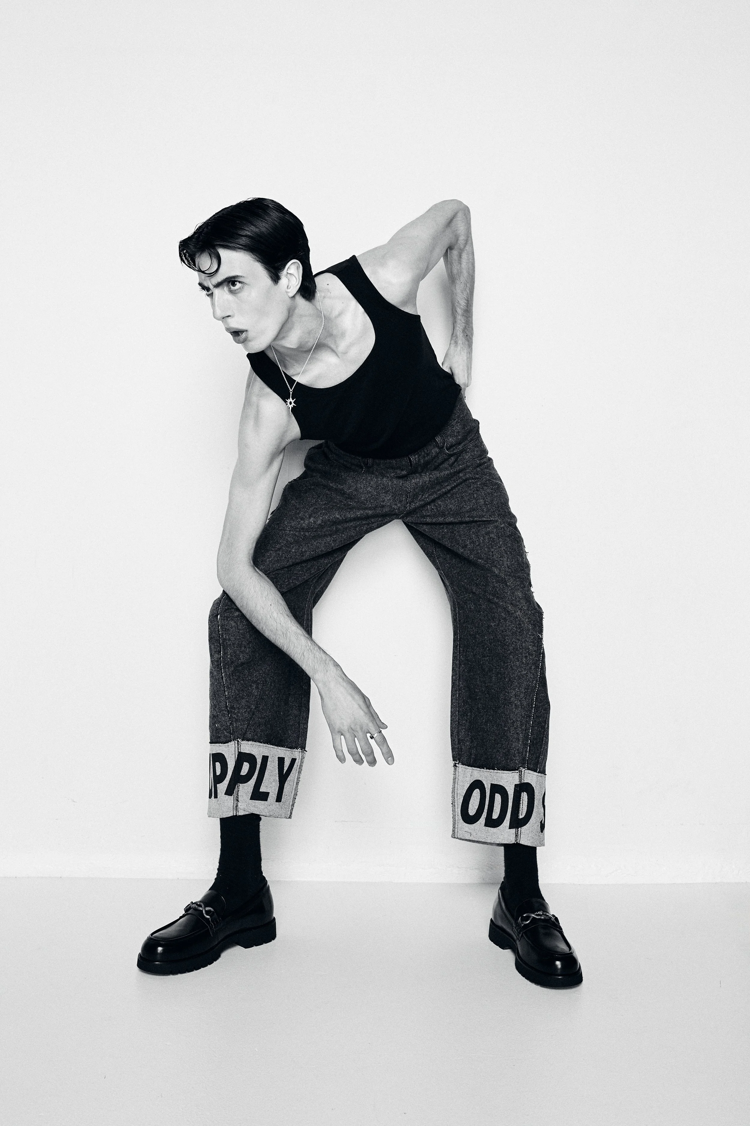Black and white photo of a person with dark hair in an expressive pose, wearing a black tank top, dark trousers with 'HAPPY' and 'ODD' written on the cuffs, and black shoes, against a plain white background.