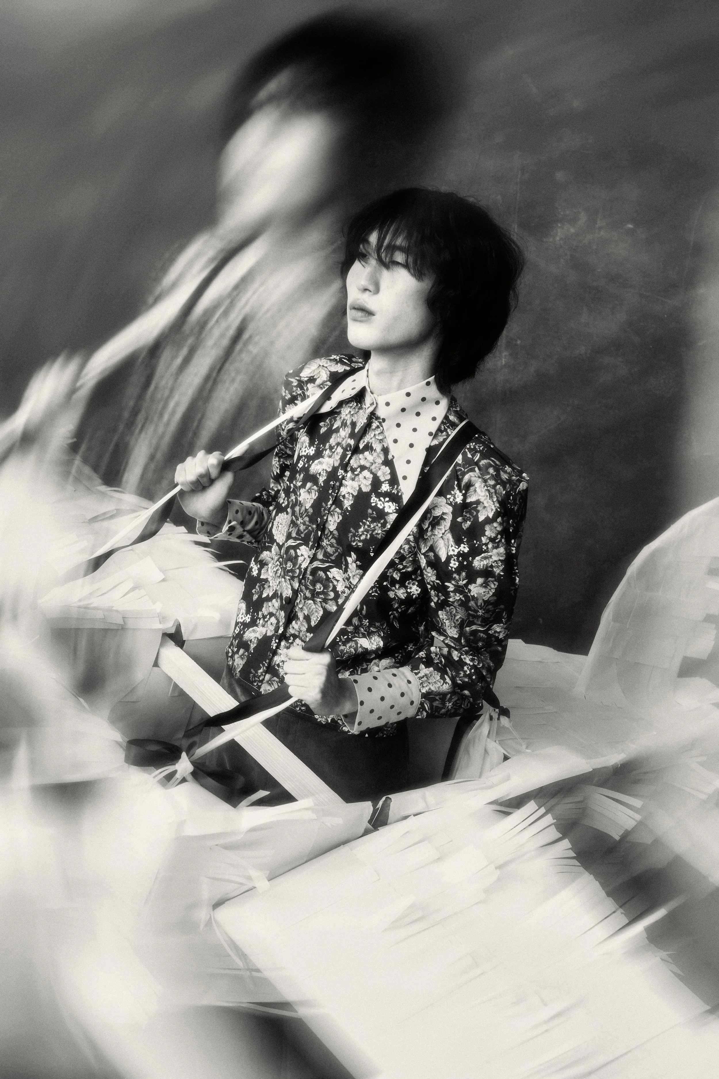 A woman with dark, wavy hair wearing a floral jacket over a polka-dot shirt, holding an umbrella, seated in a scene with blurred paper or fabric around her, in black and white.