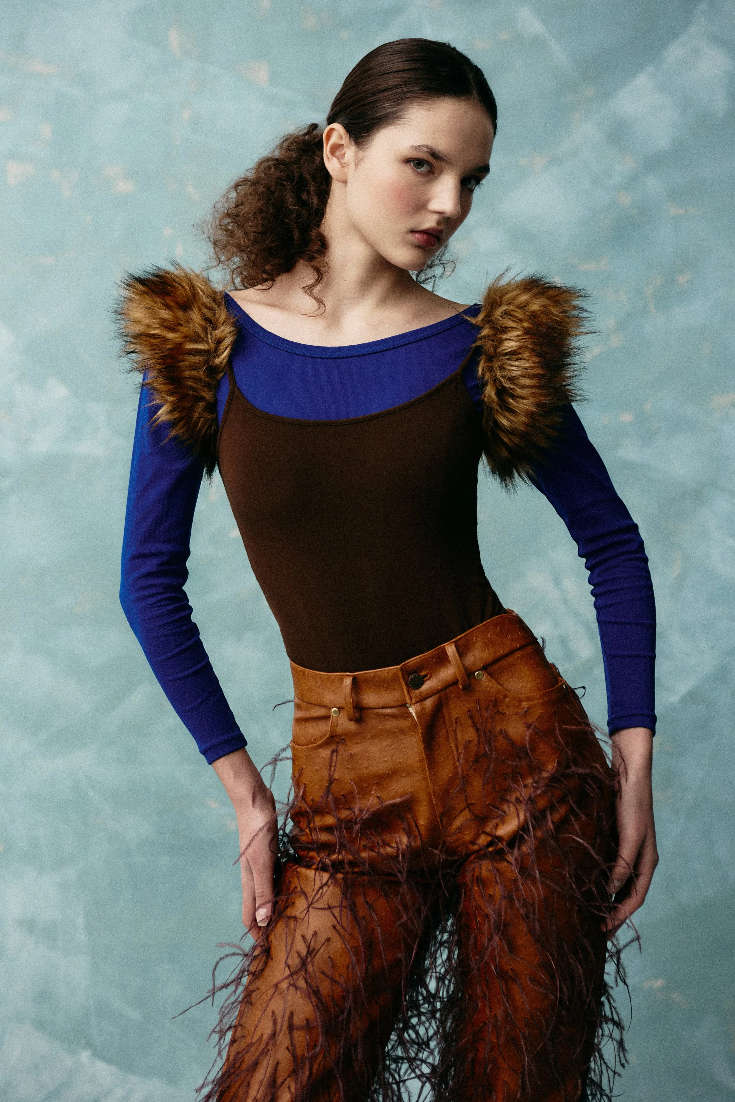 A woman with brown hair tied in a ponytail, wearing a long-sleeved blue shirt with a brown top layered over it, adorned with faux fur shoulder accents, and high-waisted brown leather pants with feather details.