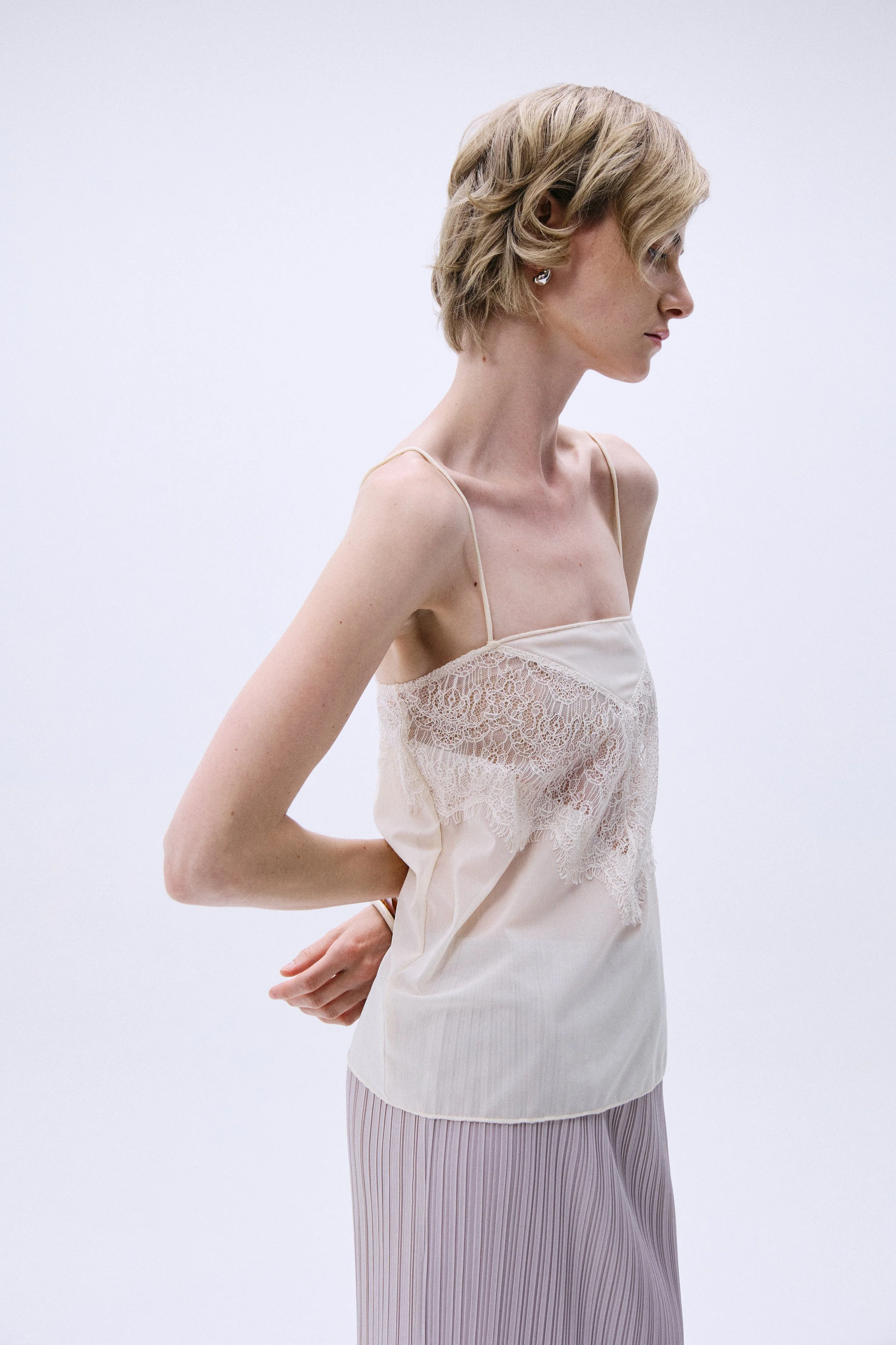 A woman with short blonde hair and earrings, wearing a silky beige camisole with lace details and a matching skirt with vertical purple stripes, standing against a plain light background in a side profile.