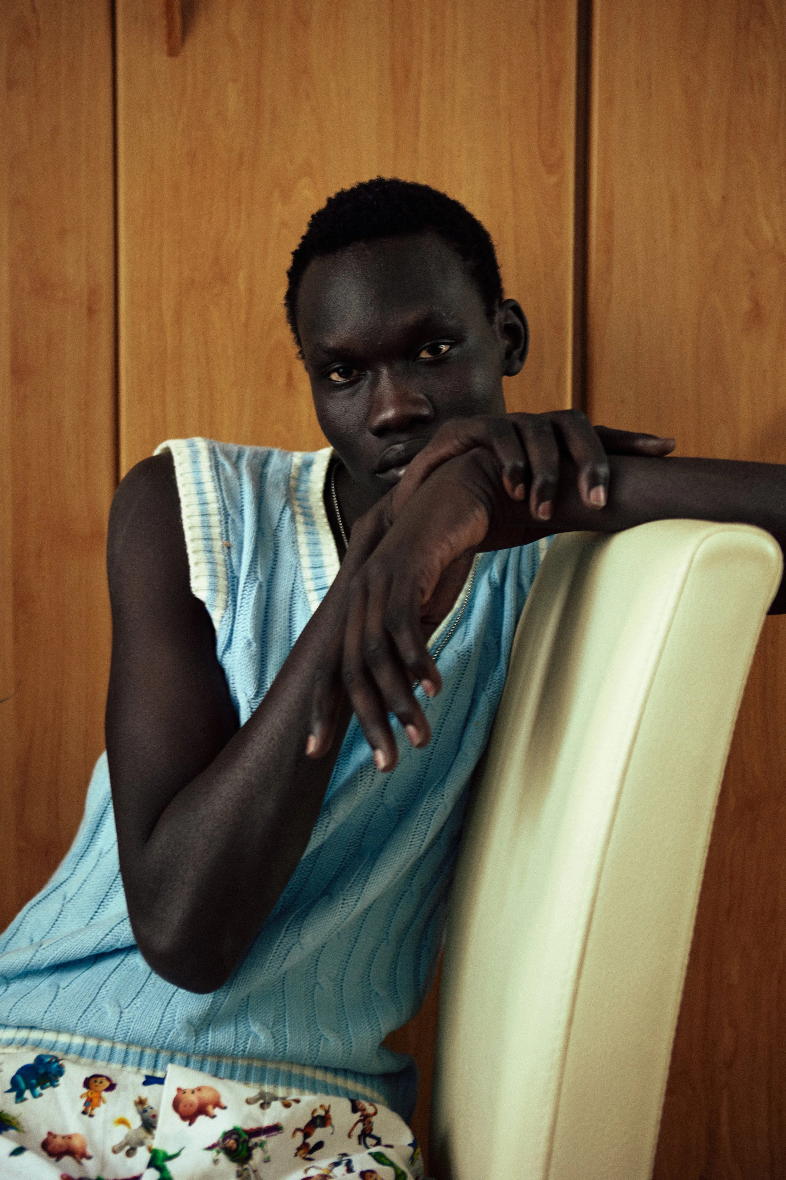 Young person with dark skin and short hair sitting in front of a wooden wall, wearing a light blue striped sleeveless shirt and with their chin resting on folded arms, looking at the camera.