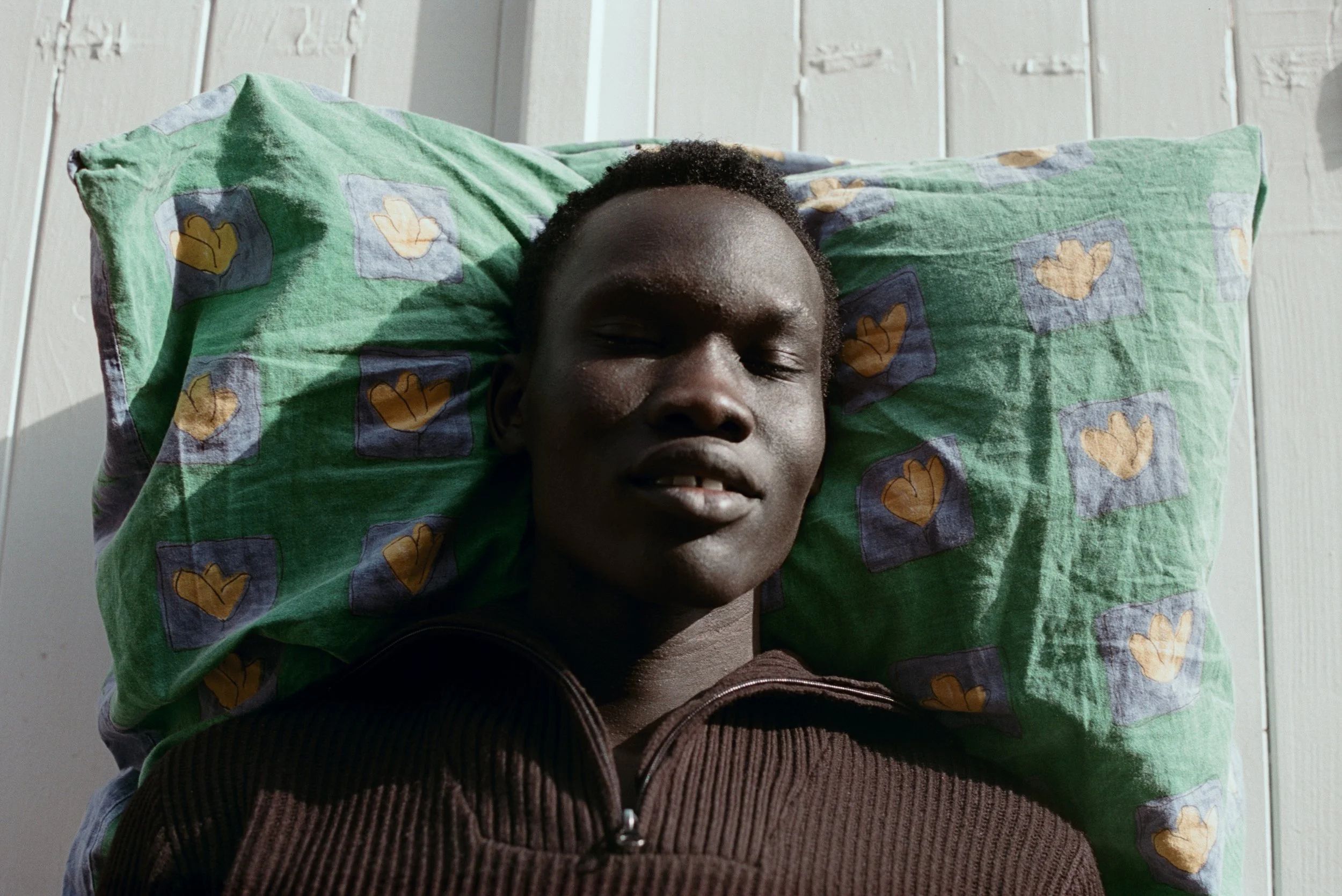 A person lying on a green pillow with a pattern of yellow and gray shapes, resting against a white wall with vertical paneling.