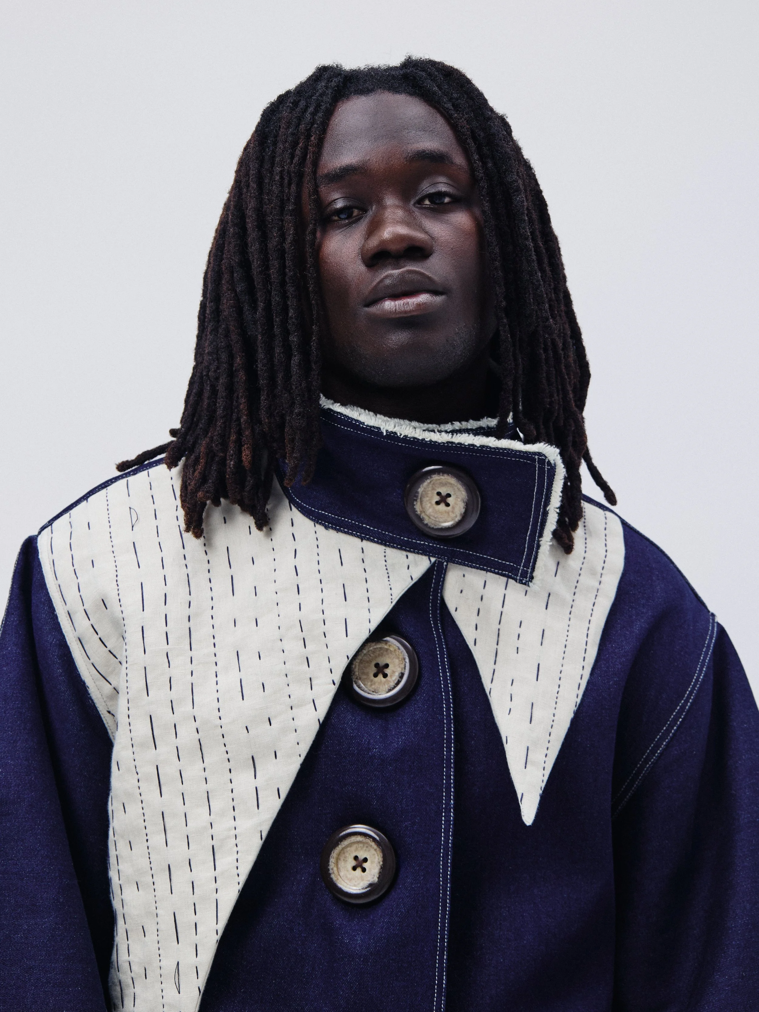 Portrait of a person with dark skin, long dreadlocks, wearing a stylish jacket with large buttons and a high collar. The background is plain and light-colored.