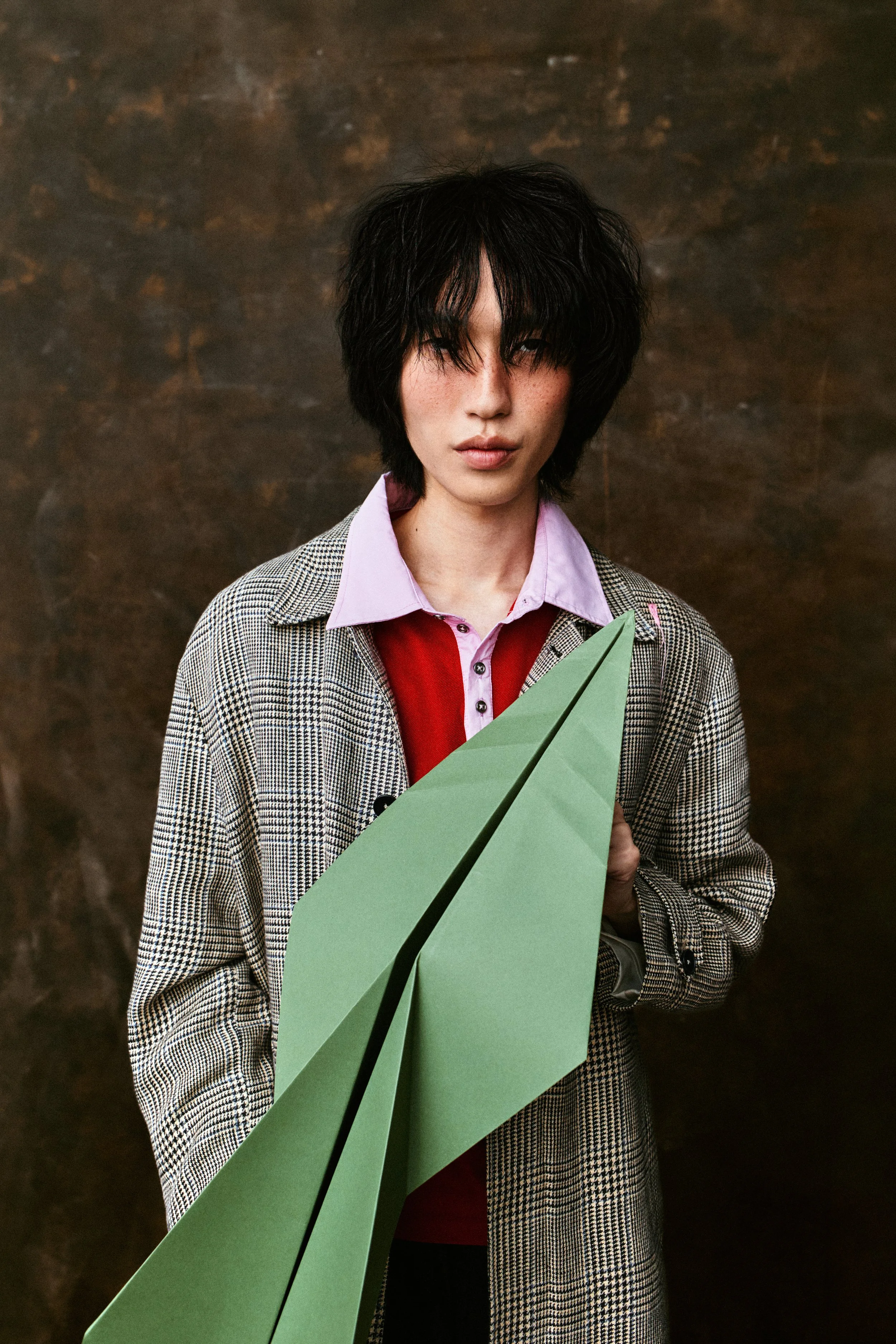 A person with black, messy hair and freckles wearing a plaid blazer over a pink and red shirt, holding a green paper airplane, standing in front of a dark brown textured background.