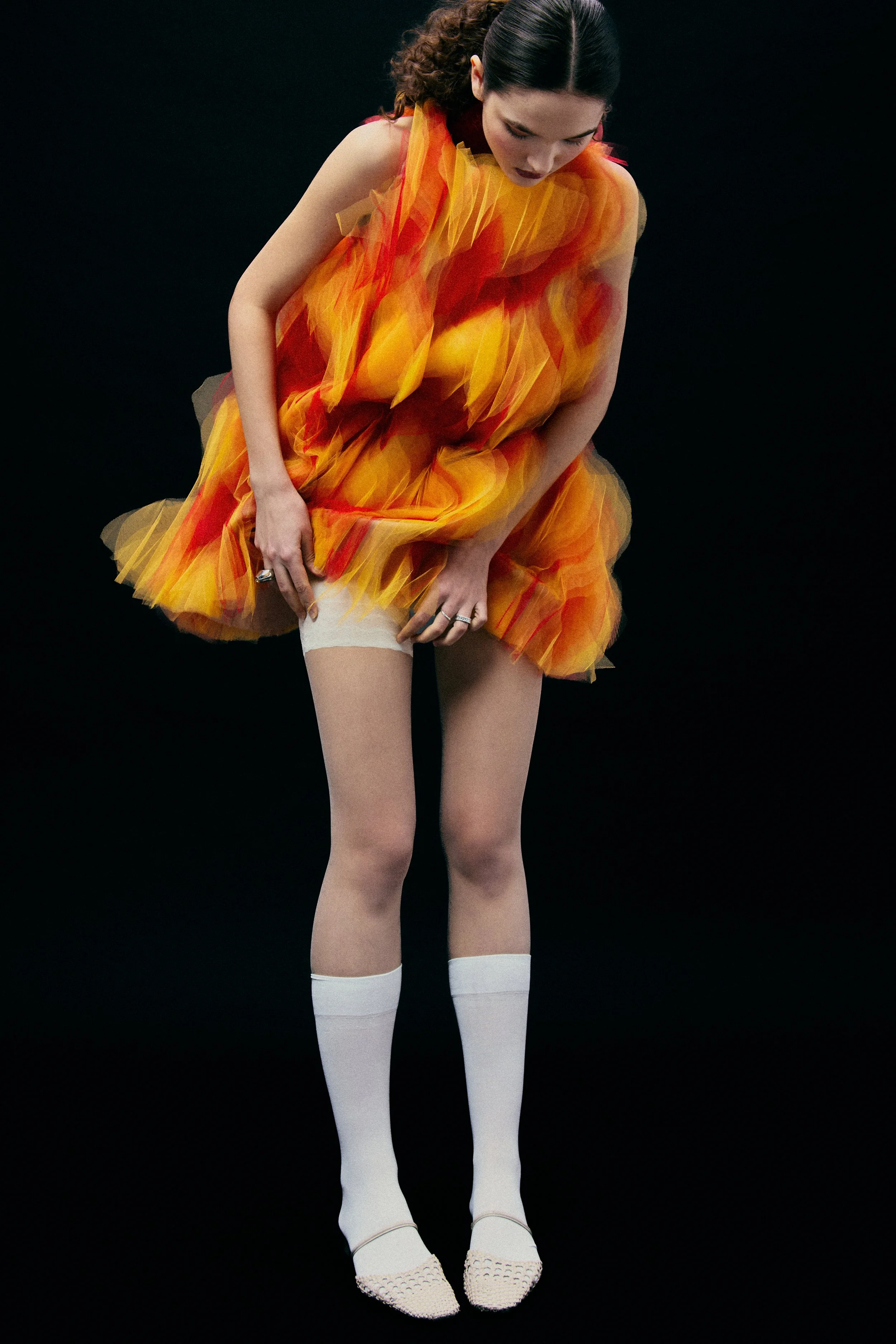 A woman wearing a colorful, orange-yellow tulle dress with white knee-high socks and beige shoes, standing against a black background.