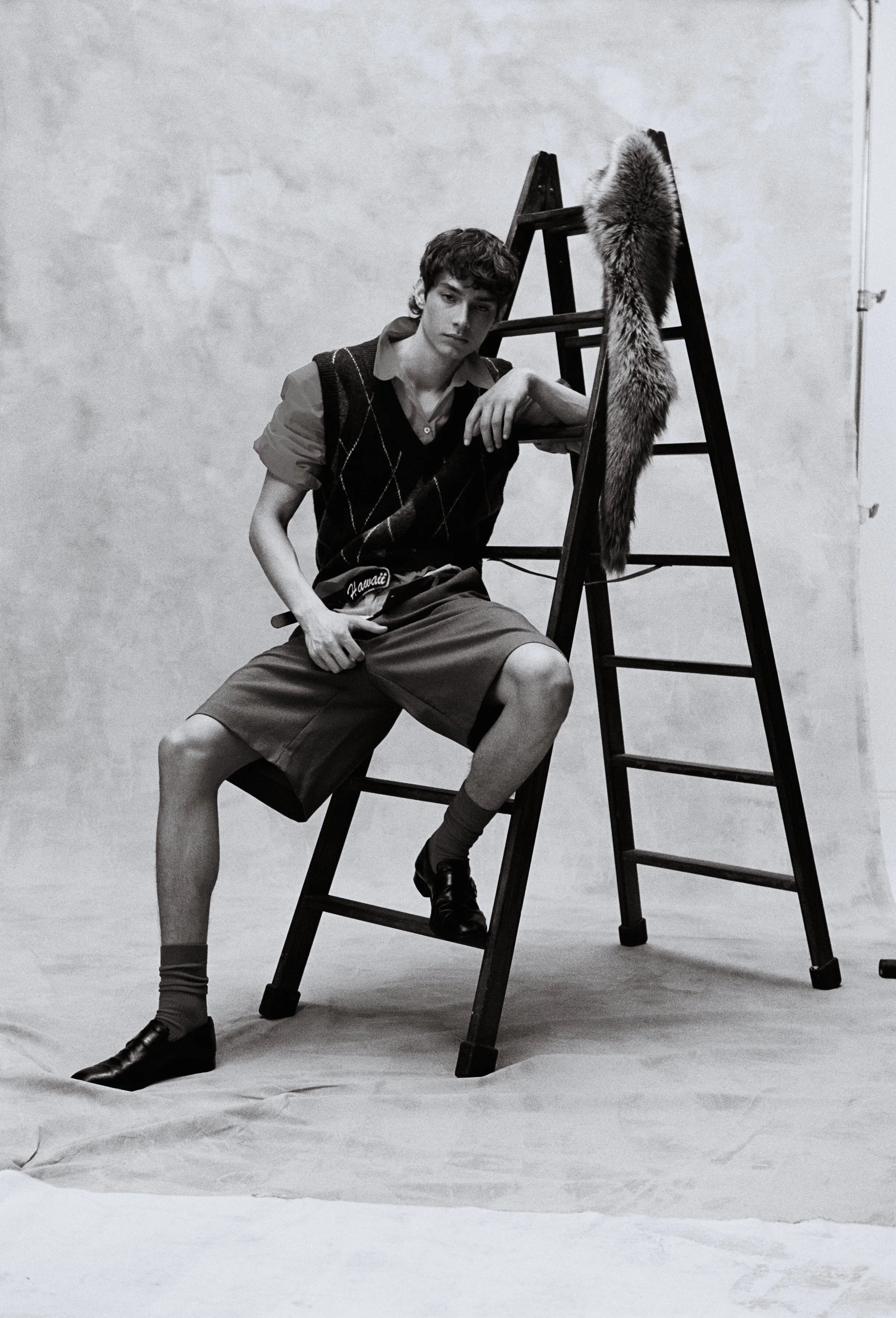 A young man in vintage style clothing, including a sweater vest, shorts, and pointed shoes, poses next to a ladder with a fur stole hanging from it. The photo is in black and white.