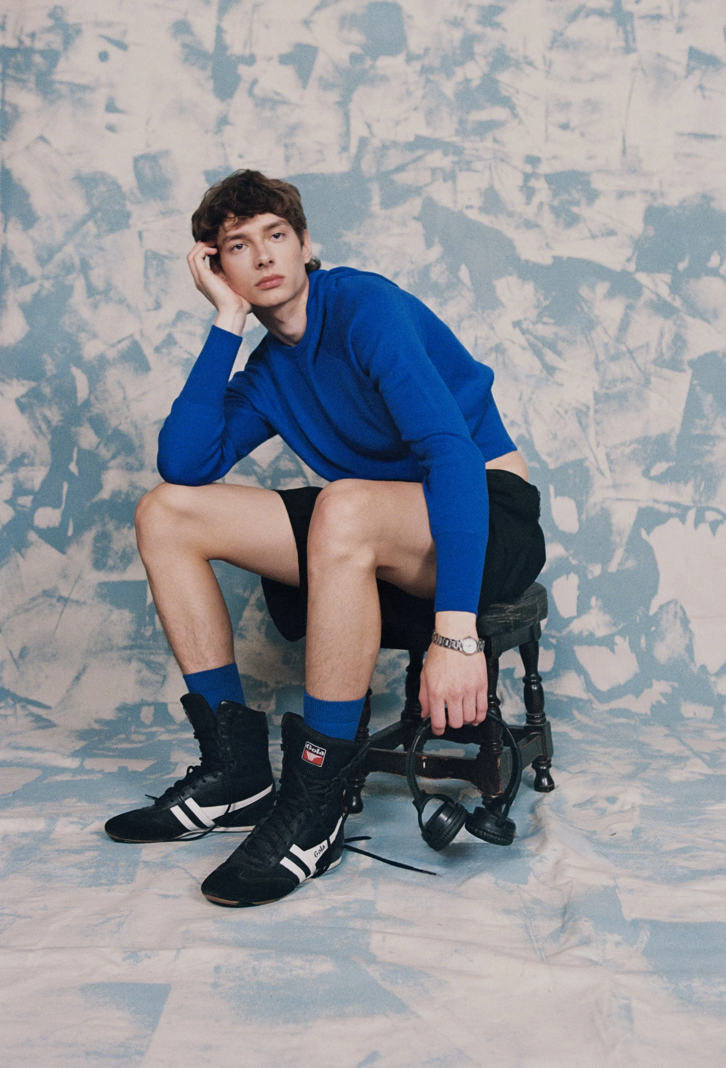 Young man in a blue long-sleeve shirt, black shorts, and black sneakers sitting on a small chair against a textured blue and white background. He rests his head on his hand, looking thoughtfully at the camera.