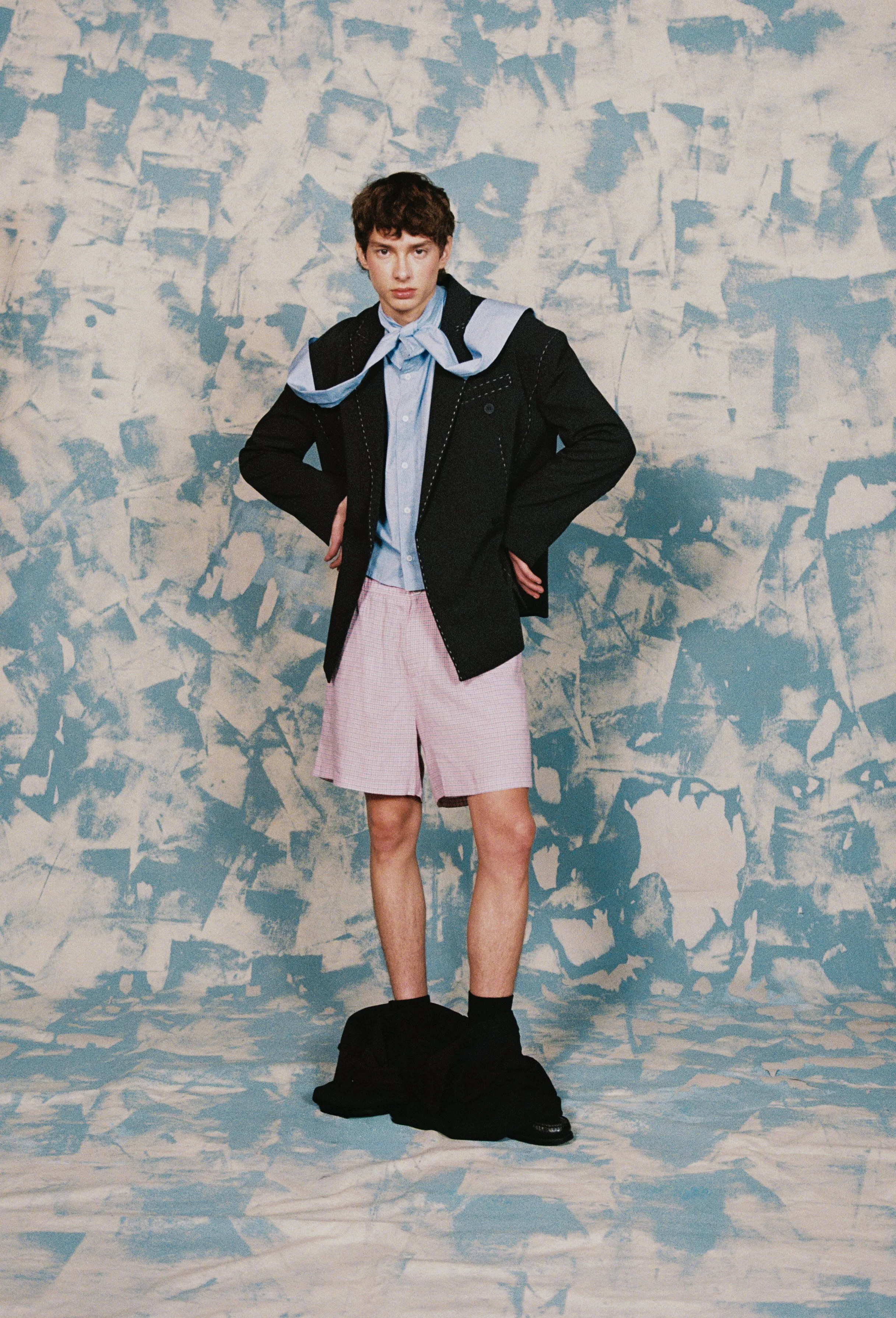 A young person with short curly hair standing in front of a textured blue and white wall. They are wearing a black jacket, a blue shirt with a scarf-like collar, pink shorts, and black shoes with black socks. The person's hands are on their hips, and