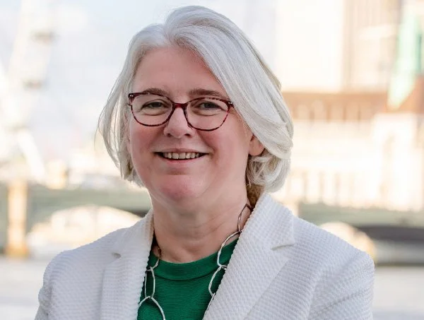 A middle-aged woman with gray hair, glasses, wearing a white blazer and a green top, smiling outdoors with a cityscape and water in the background.