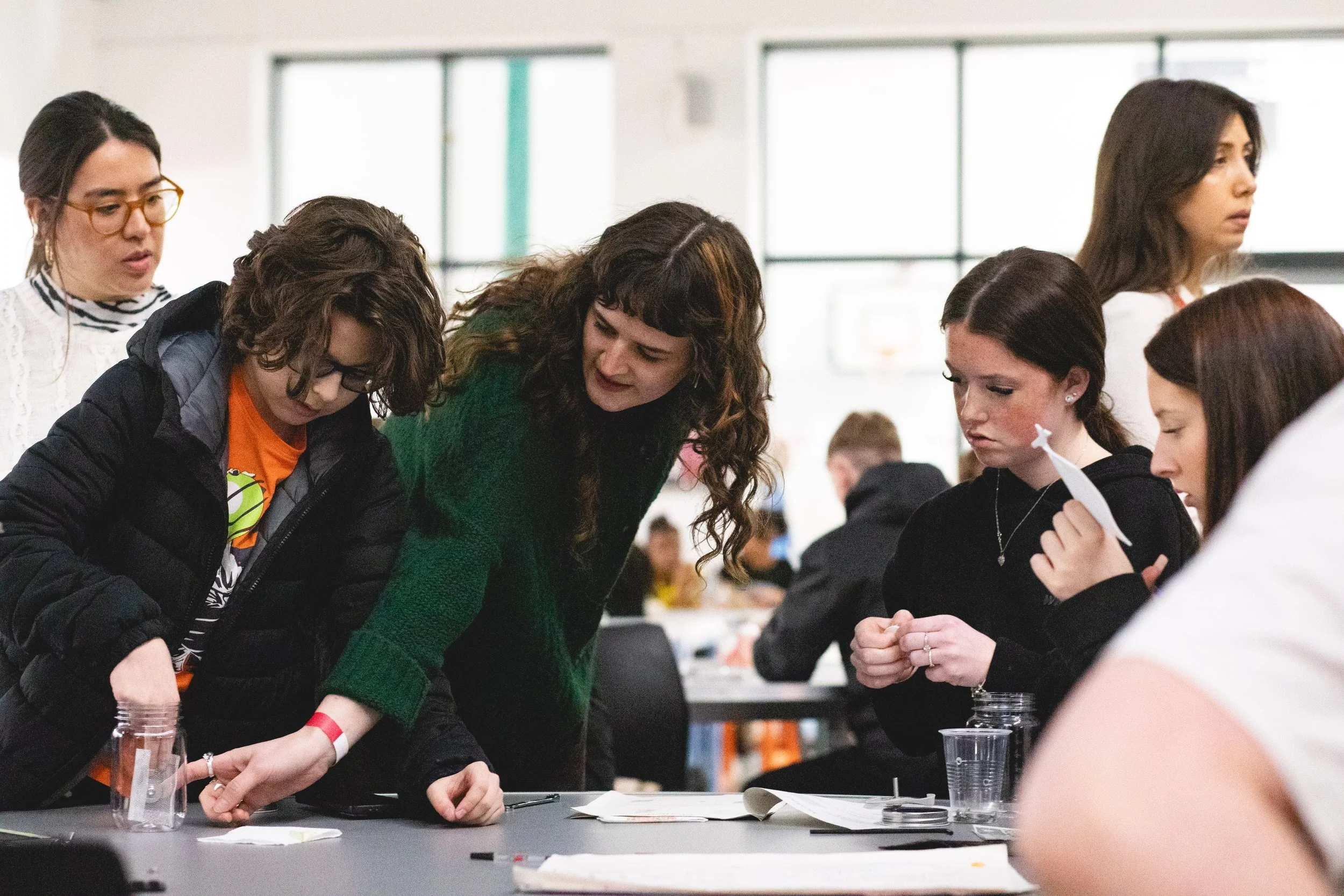 A group of women gathered around a table engaged in a craft or science activity, with some holding clear plastic cups and others focusing on their work.