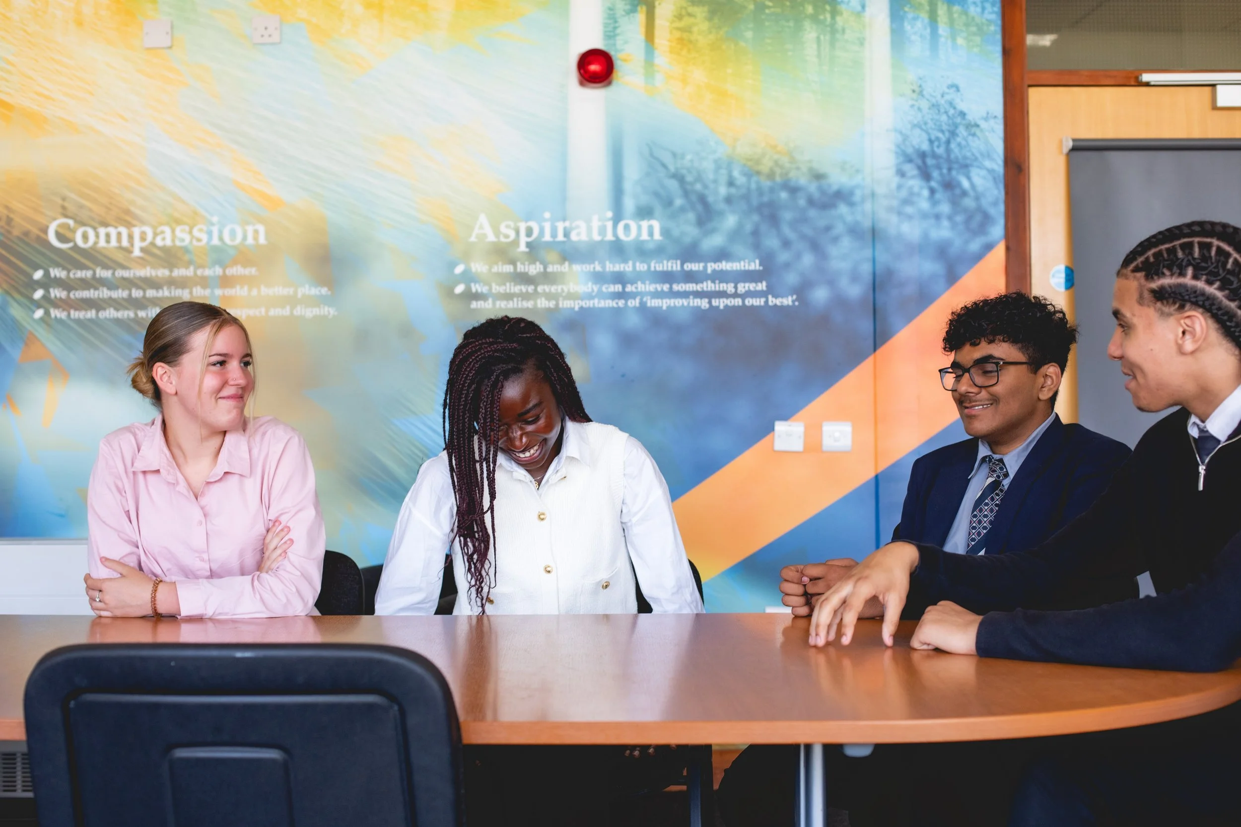 Four diverse young adults sitting at a conference table in a brightly colored room, smiling and engaged in conversation. A large mural with text about compassion and aspiration decorates the wall behind them.
