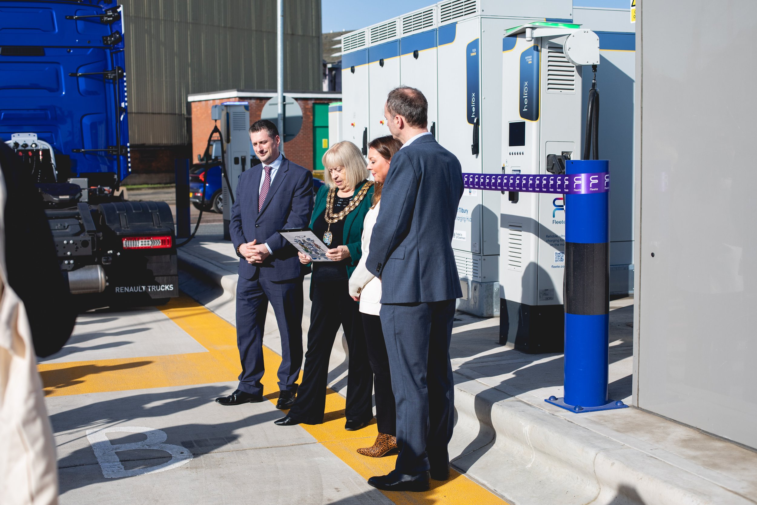 A group of four business professionals, three men and one woman, standing outside next to electric vehicle charging stations and a truck, engaging in a discussion while holding documents.
