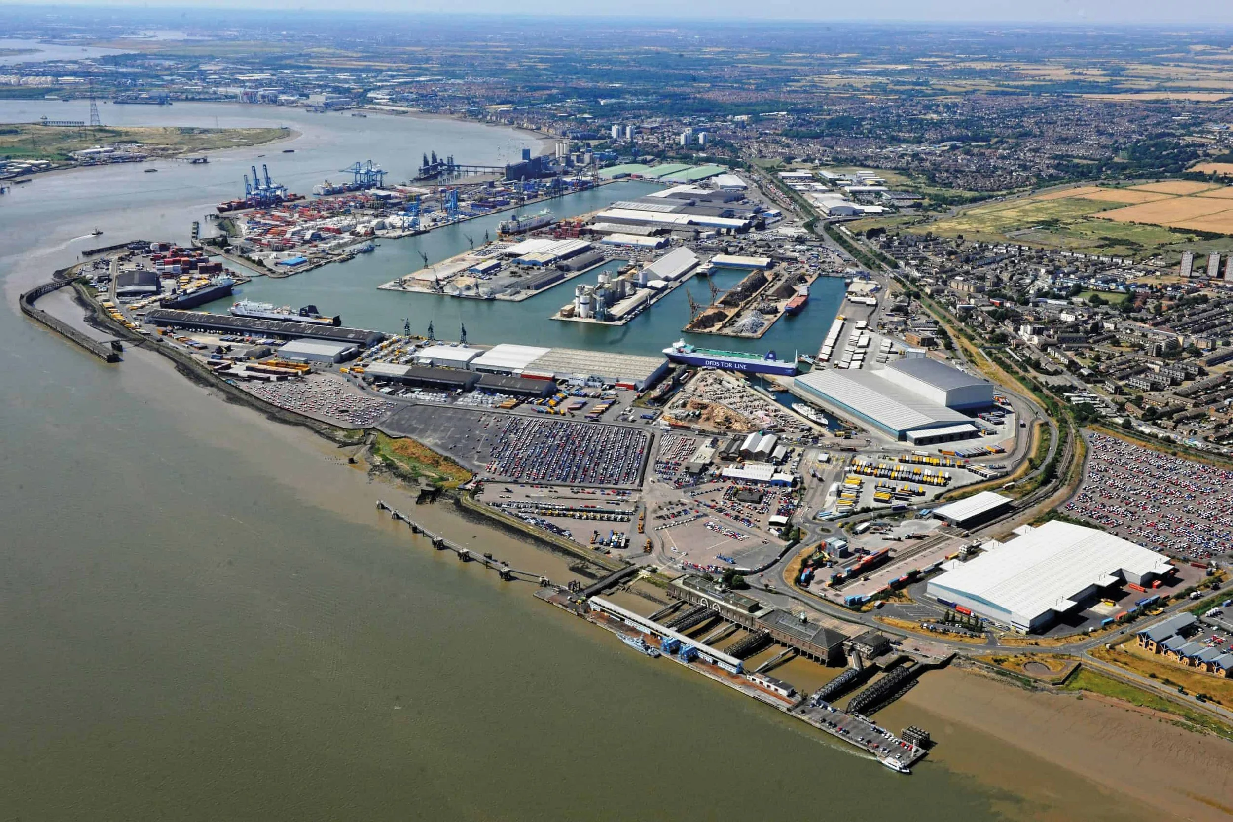 An aerial view of a large port with shipping containers, cranes, warehouses, and a ferry docked at the terminal along a river, with a city and farmland in the background.