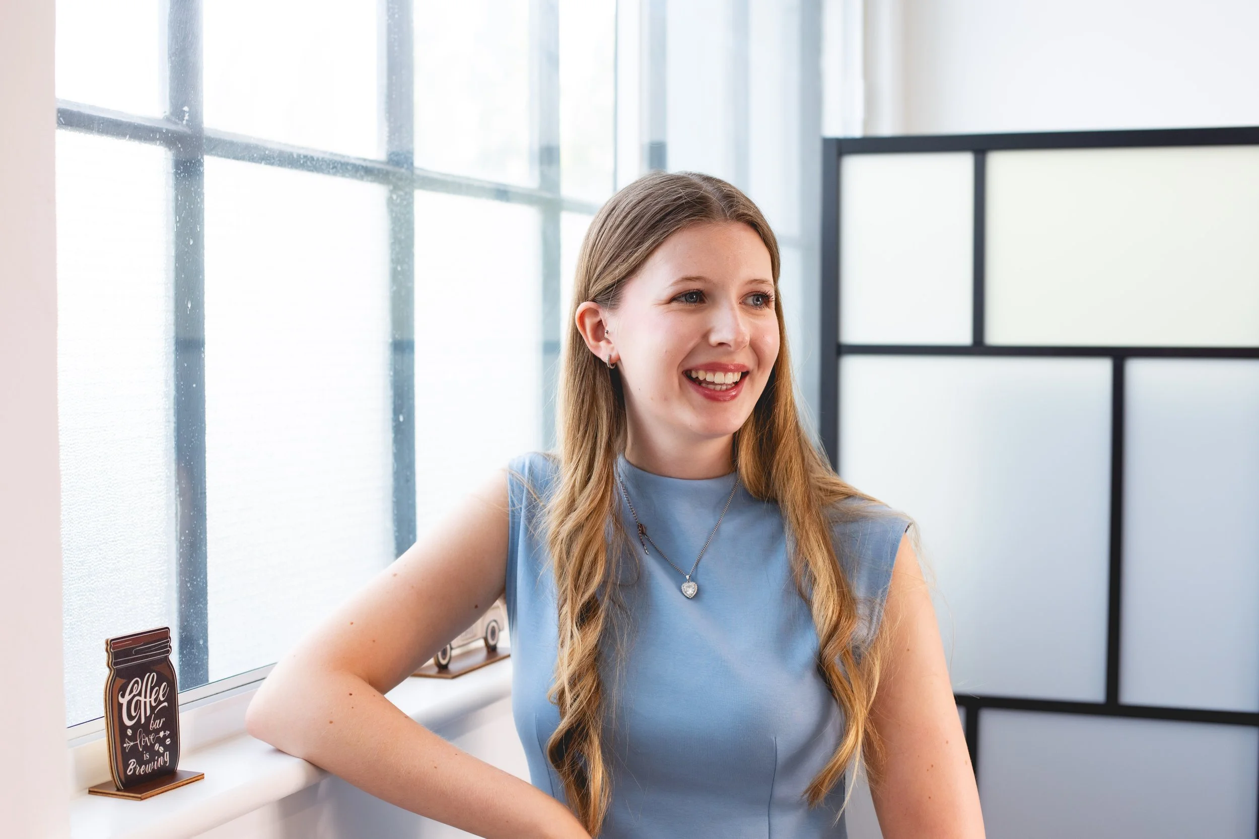 A young woman with long, wavy blonde hair wearing a sleeveless, light blue top and jewelry, smiling and sitting by a window with natural light, in an indoor setting.